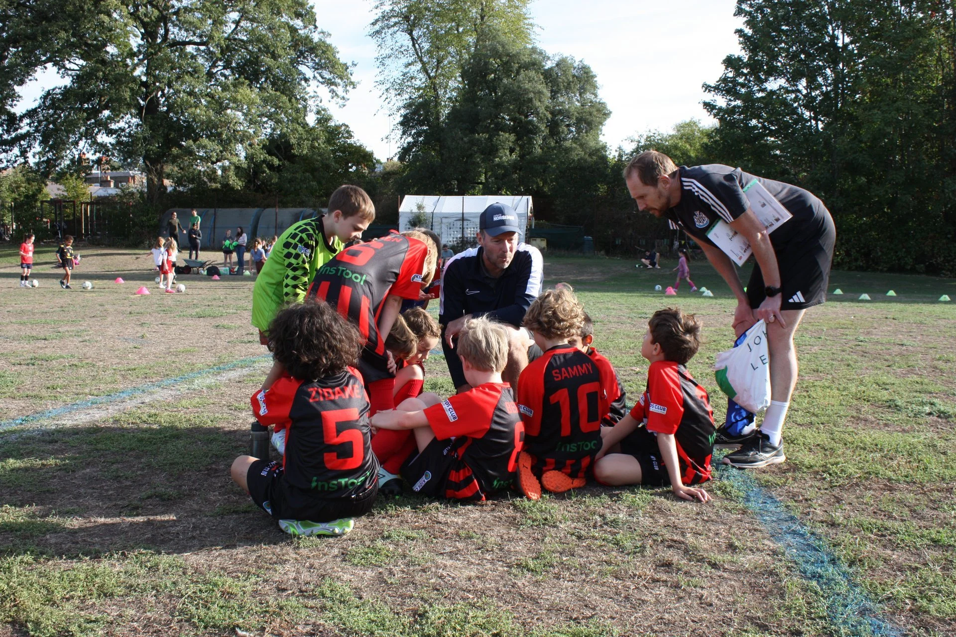 A youth soccer team sitting in a huddle on the field while their coaches give instructions. The players wear red and black uniforms with number and name on the back. In the background, young children practice soccer drills, and there are large trees and a greenhouse or shed.