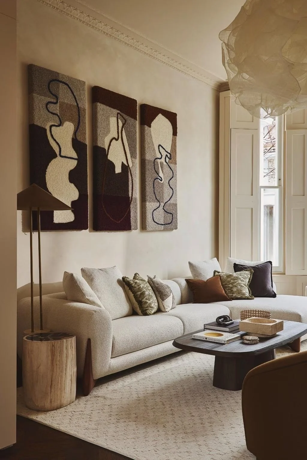 A living room with a beige sofa decorated with various patterned pillows, a dark wooden coffee table holding books, a bowl, and a pair of headphones. To the left, a tall floor lamp stands beside a wooden side table. The wall behind the sofa features three abstract art pieces, and a large window with white shutters lets in natural light.