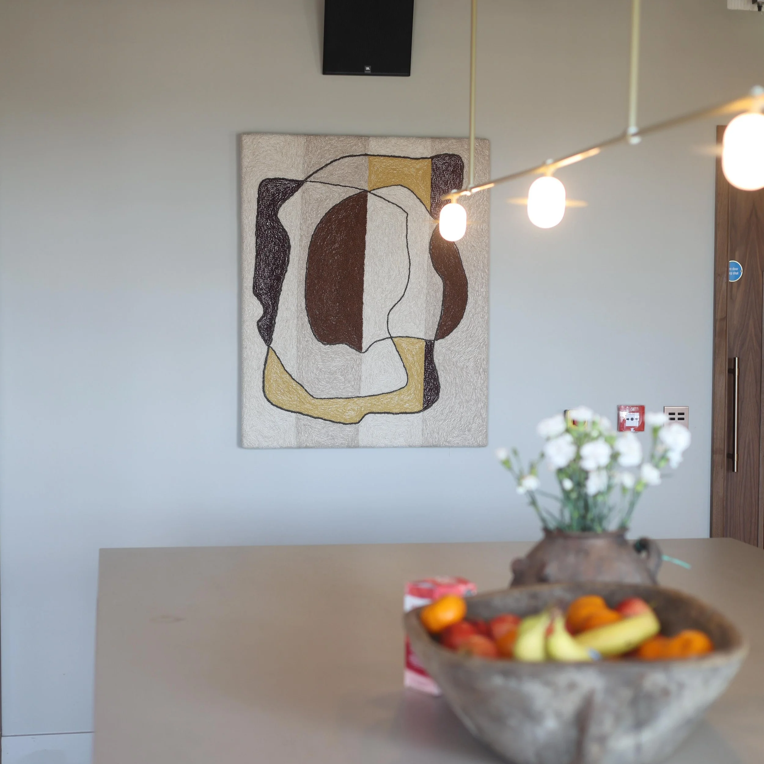 Interior of a room with a modern abstract painting on the wall and hanging lights. In the foreground, a blurred bowl of colorful candies or fruit is visible.