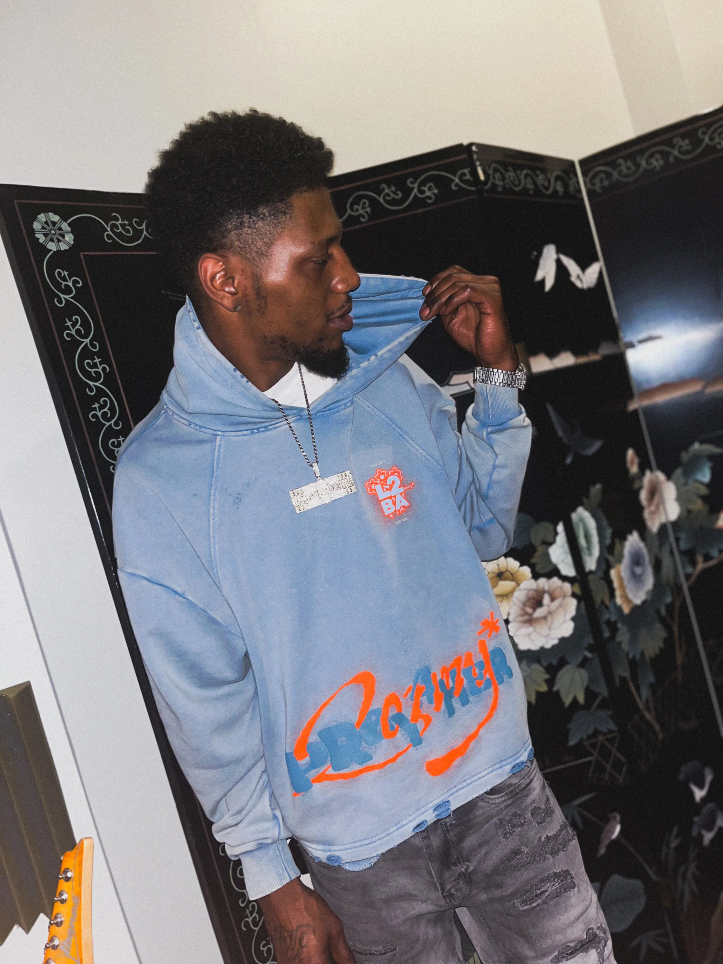 A young man with curly hair and a beard wearing a Blue Pray 4 Her HARMMONEY* hoodie. He is wearing a watch and a chain necklace, standing in a room with floral and decorative wall art.