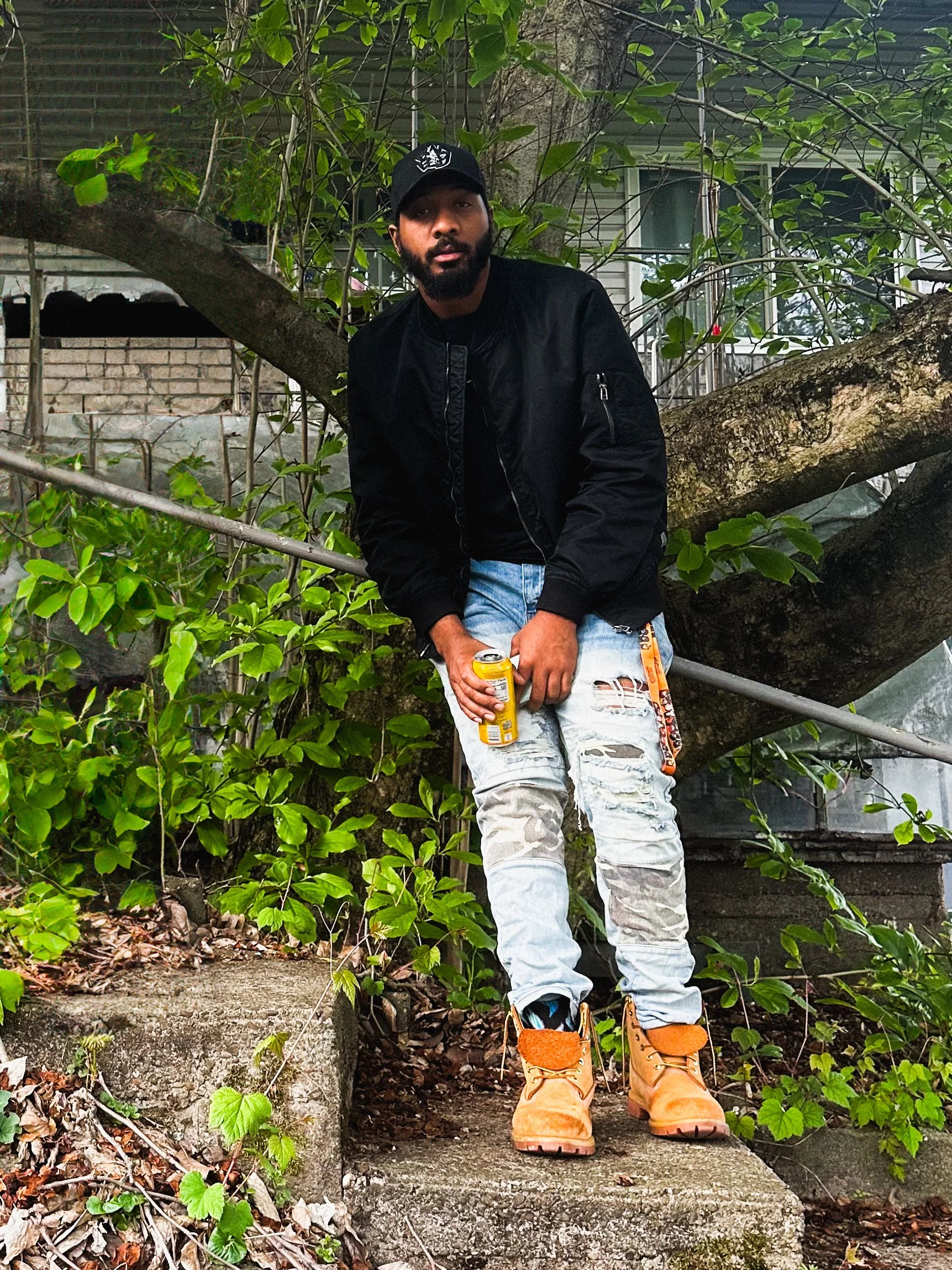 HARMMONEY artist NeedEvryStak standing on steps in front of overgrown house with a tree