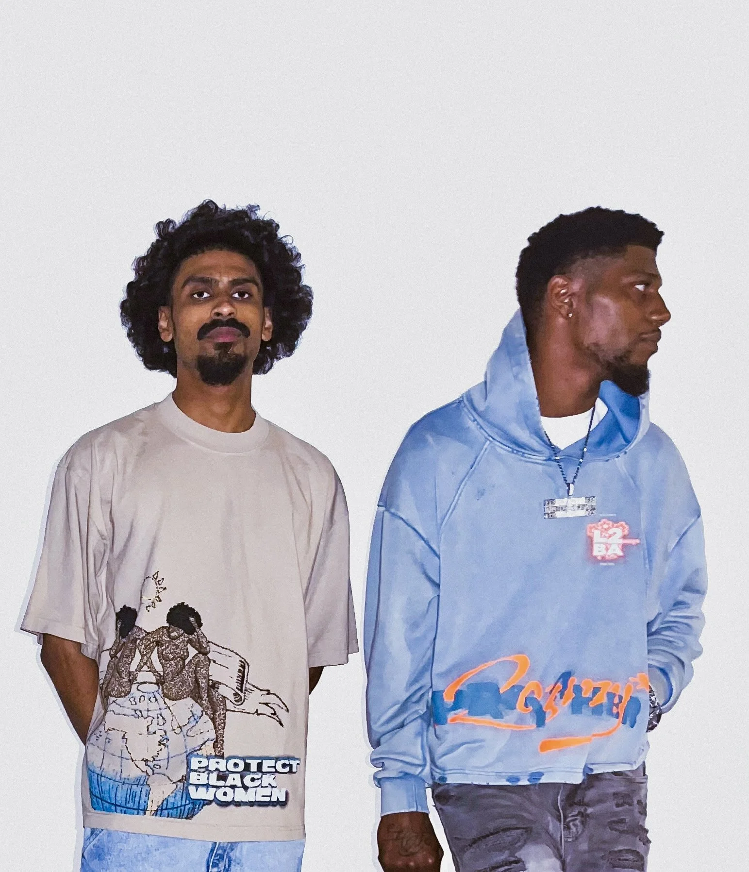 Two young men standing against a plain white background. The man on the left is wearing the HARMMONEY "Protect Black Women" Tee, the man on the right is wearing a blue HARMMONEY Pray 4 Her Hoodie