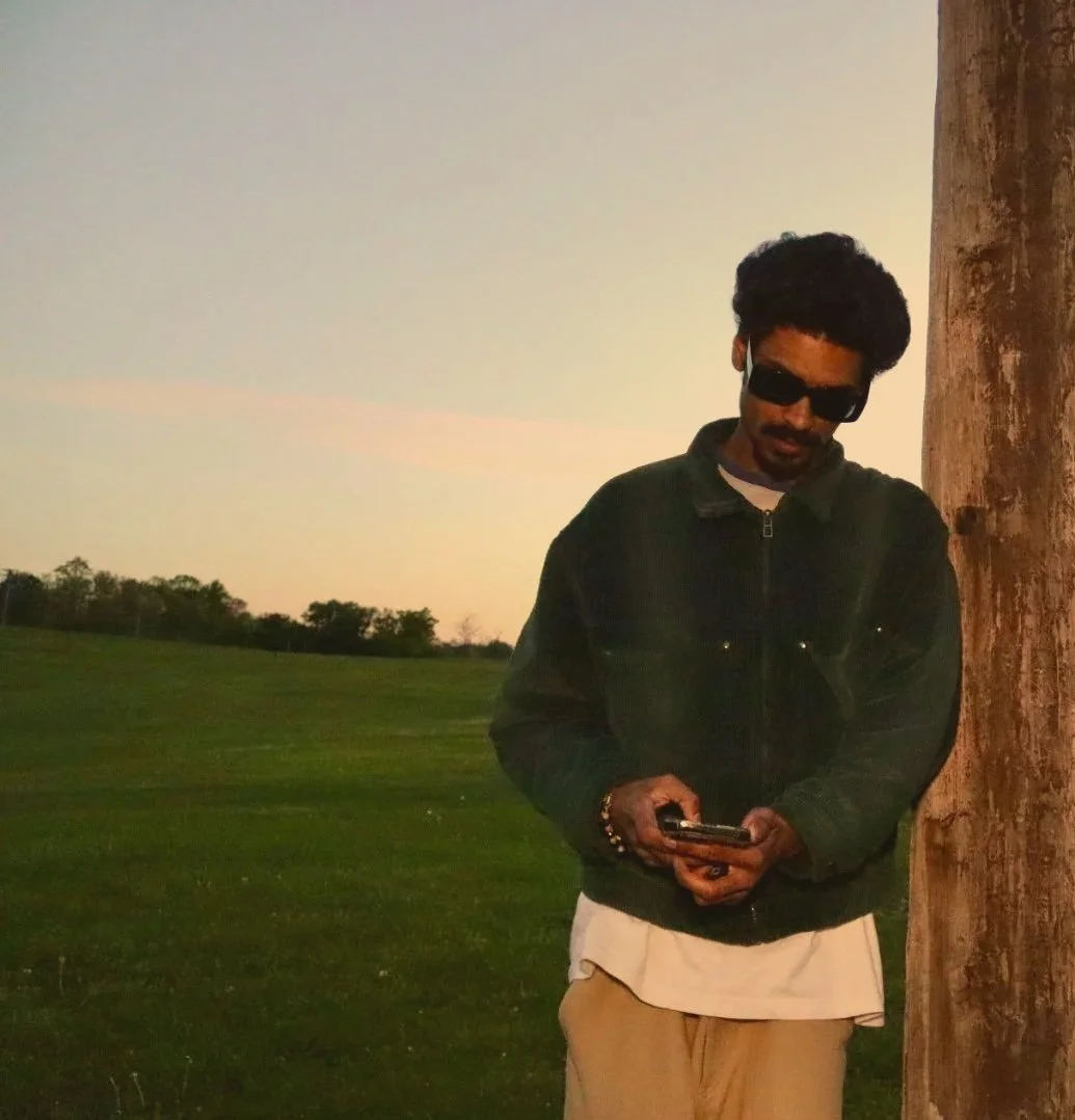 Rapper, 2onny*, standing outdoors at sunset by a tree, looking at his phone, wearing a teal jacket, white shirt, and beige pants, with a grassy field and trees in the background.