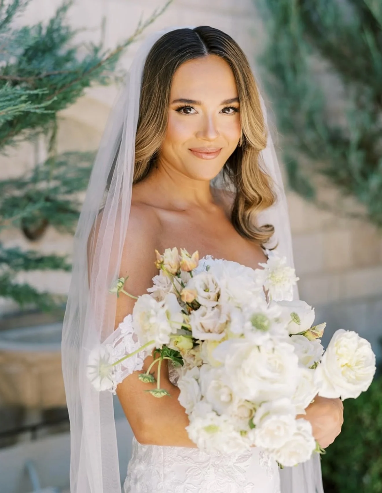 A woman in a wedding dress holding a bouquet of white flowers, with a sheer veil and outdoor greenery in the background.