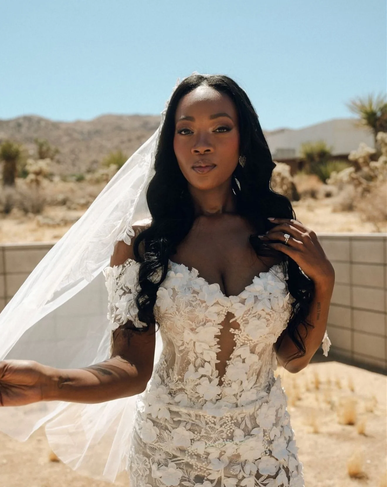A woman in a white wedding dress with floral details stands outdoors in a desert landscape with hills in the background. She has long black hair, wears earrings, a veil, and has a ring on her right hand.