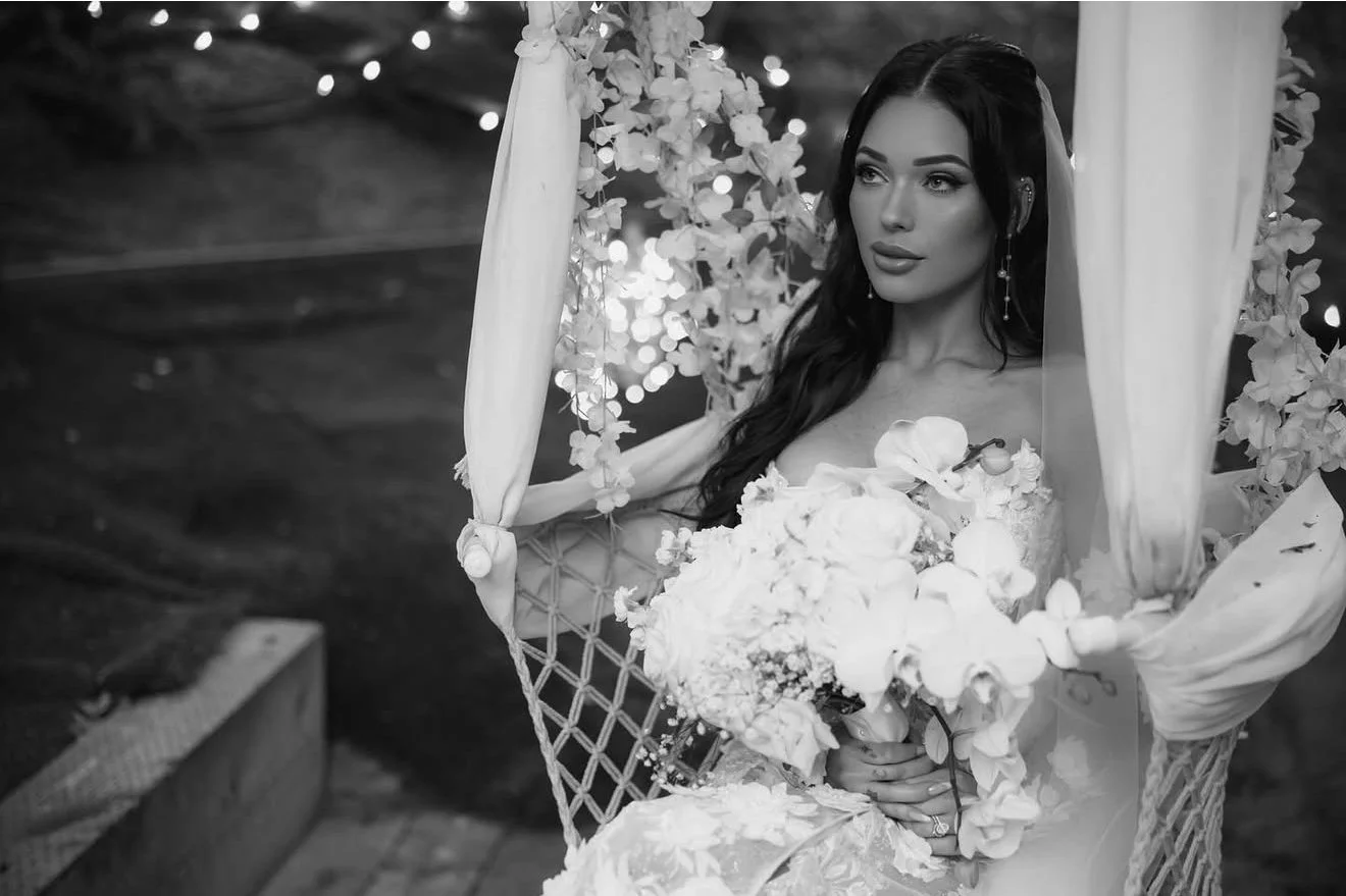 A woman in a wedding dress sitting on a decorative chair, holding a bouquet of flowers, surrounded by hanging flowers and curtains, in a setting with blurred background lights.