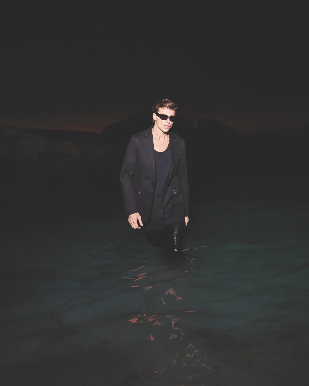 A person dressed in black, wearing sunglasses, walking through water at night with a dark sky and distant mountains in the background.