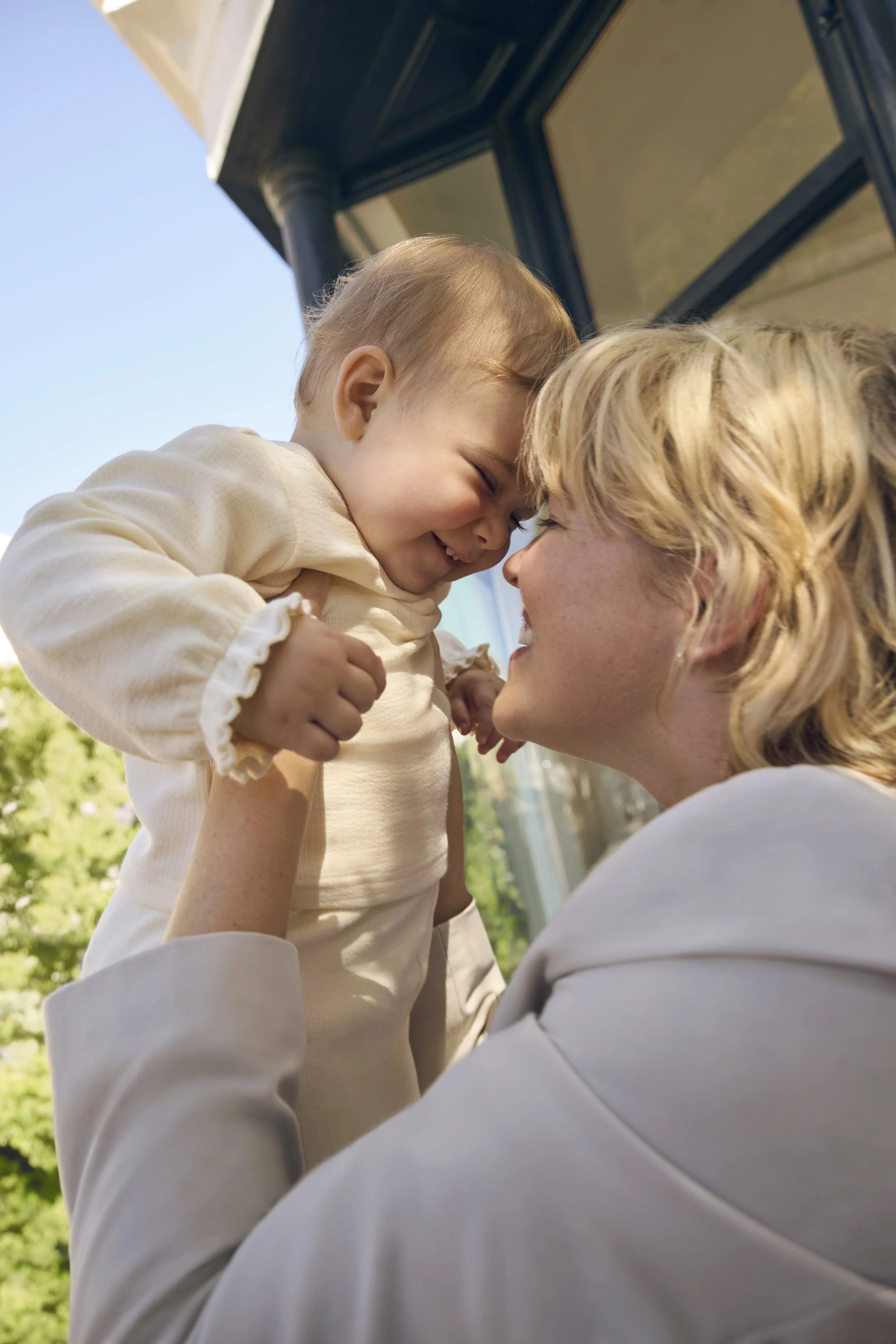 A young boy with short light hair smiling as he is held up face-to-face with an adult woman, possibly his mother, outdoors near a house with a porch and glass windows.
