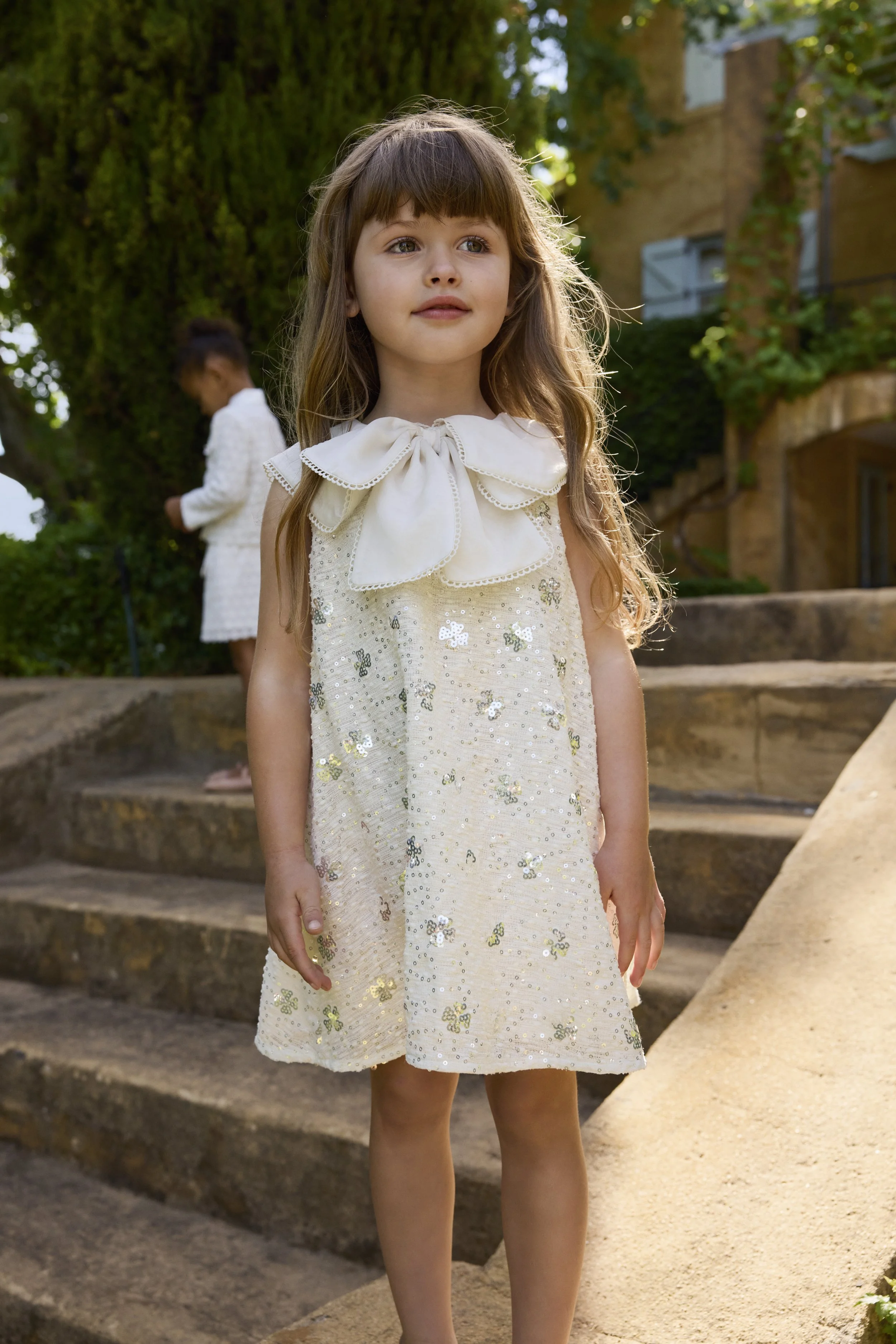A young girl with long wavy hair wearing a cream dress with sequins and floral embroidery, standing outdoors on steps with greenery and a building in the background.