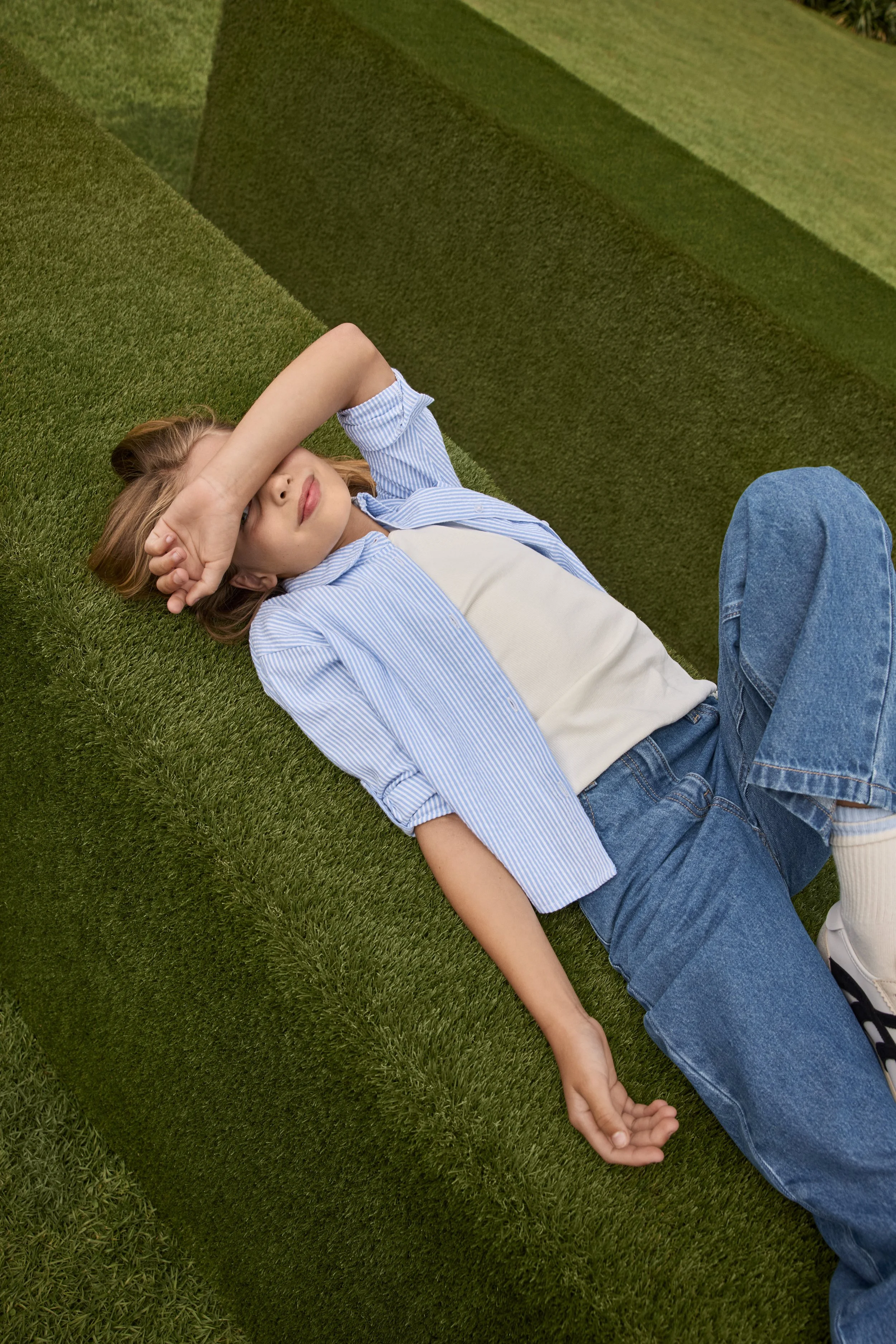 A girl with light brown hair wearing a blue striped shirt and blue jeans lying on green grass with her eyes closed, partially covering her forehead with her arm, holding hands with someone out of frame.