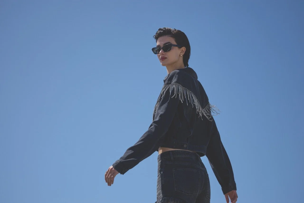A woman with dark hair and sunglasses stands outdoors against a clear blue sky, wearing a dark jacket with fringes on the shoulders and dark jeans.