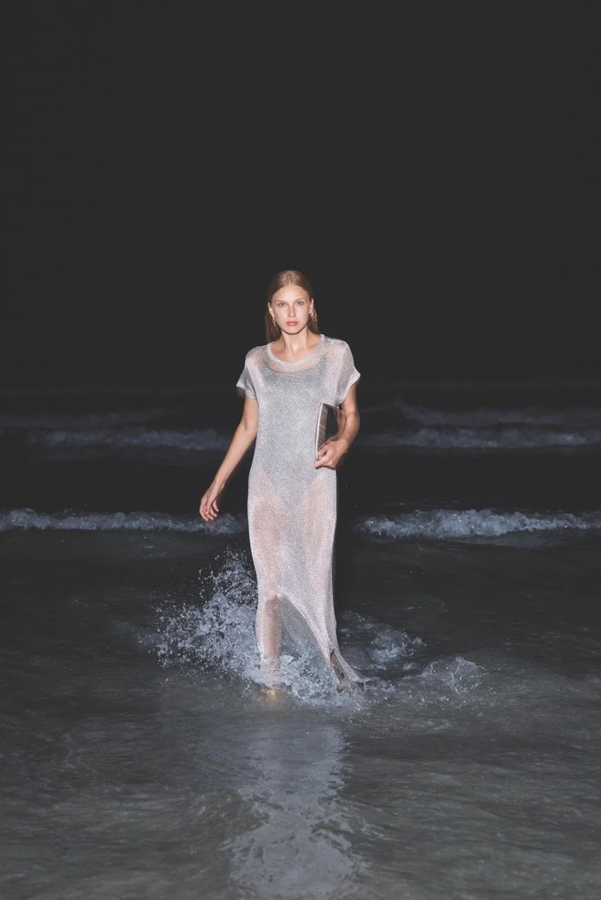 A woman in a long, silver dress walking through shallow ocean water at night, holding a book.