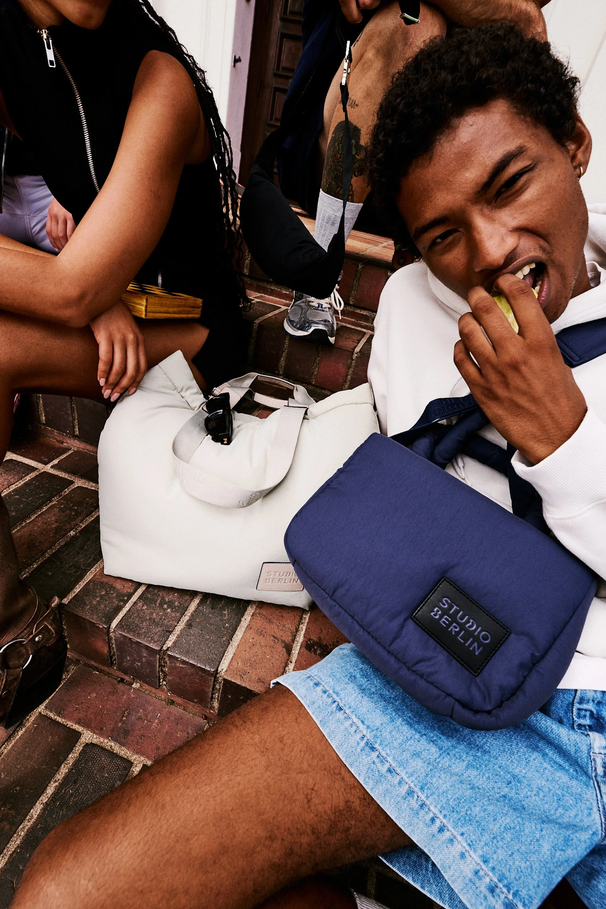 Close-up of a young man with curly hair eating an apple, sitting on the ground with a backpack and sunglasses nearby, surrounded by other people.