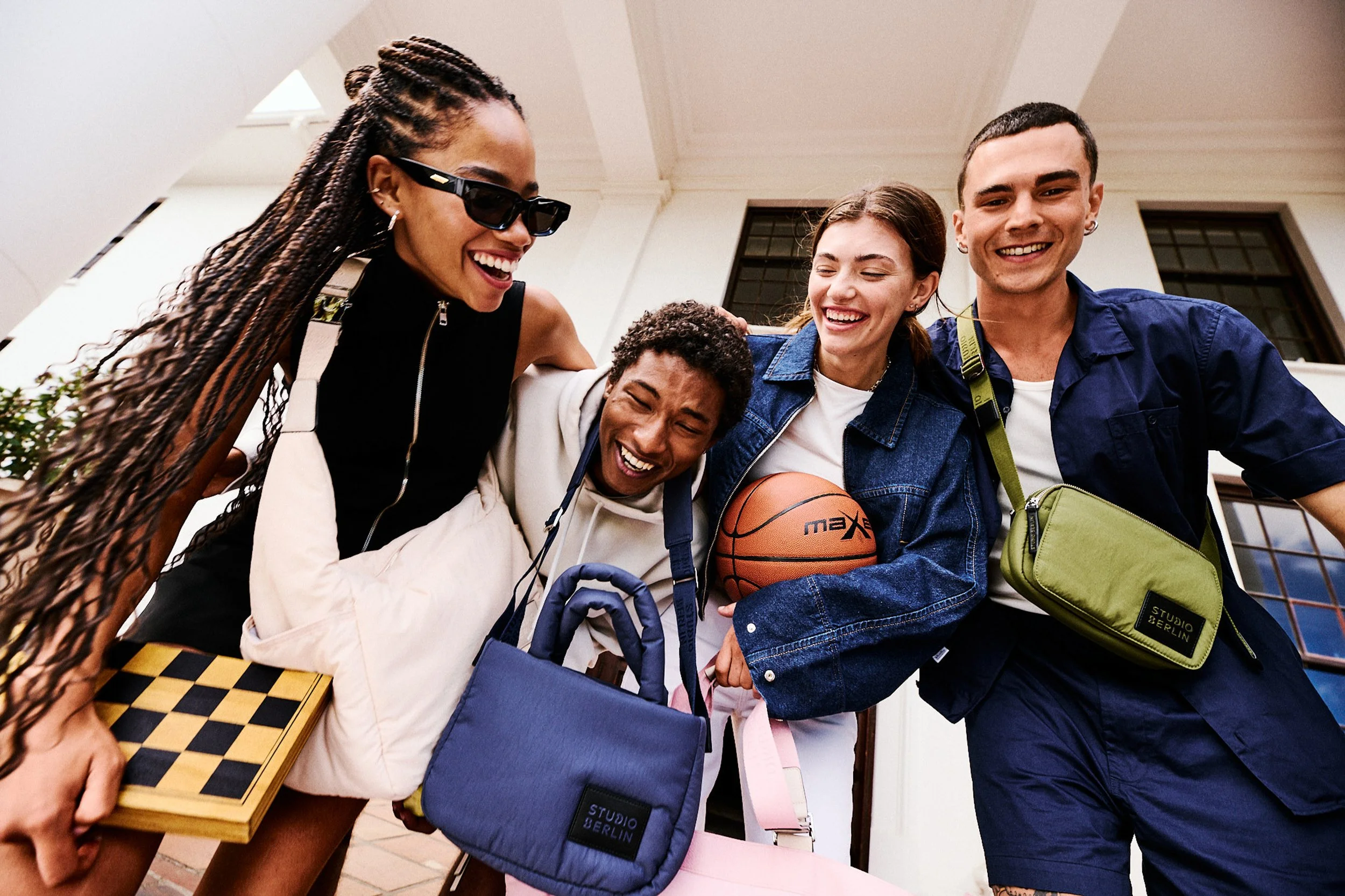 A group of five young people smiling and laughing, gathered around a table with a chessboard and a basketball, outdoors near a building with large windows.