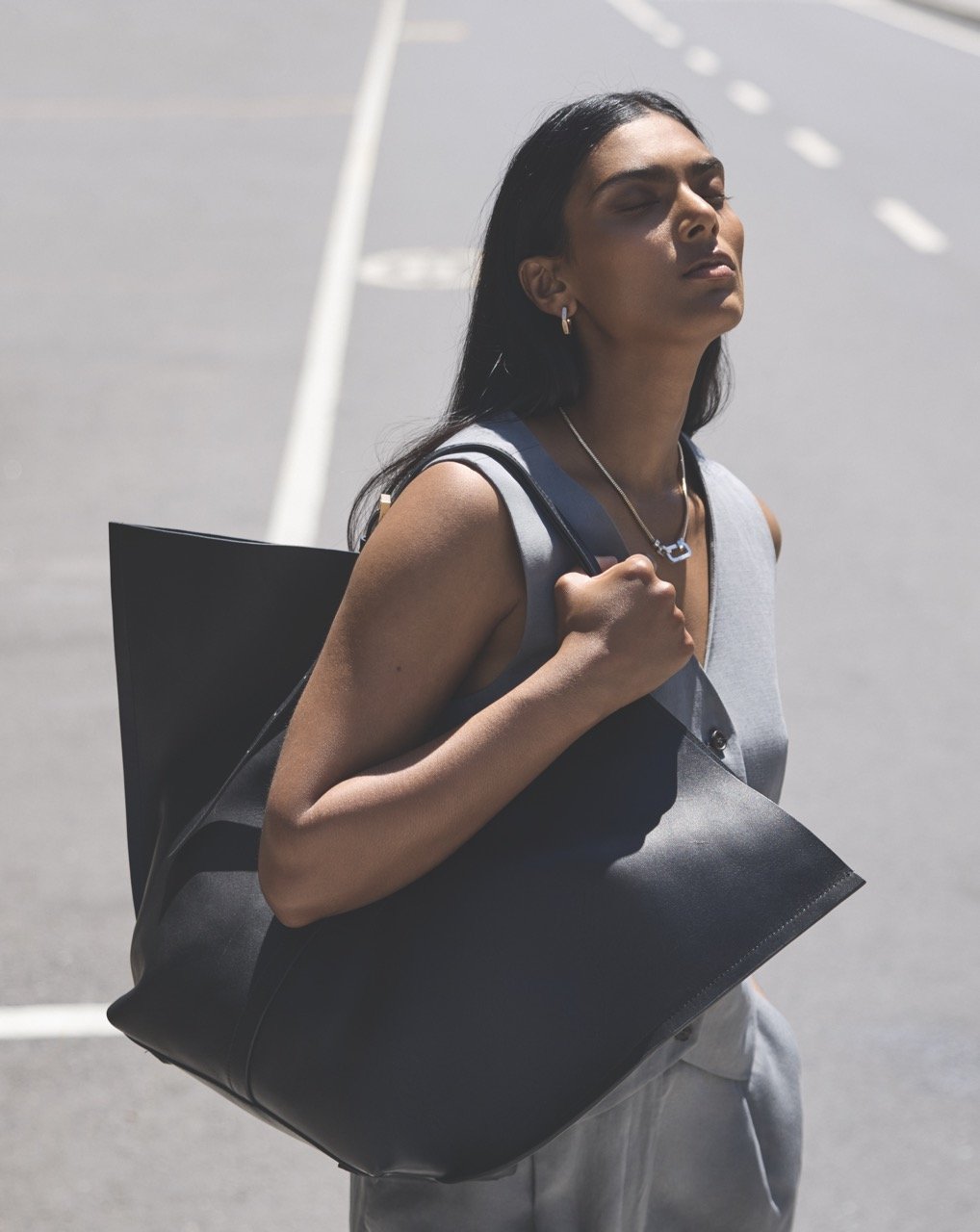 Woman with long dark hair, wearing a sleeveless top, carrying a black tote bag over her shoulder, walking on a street with her eyes closed and face tilted upward.