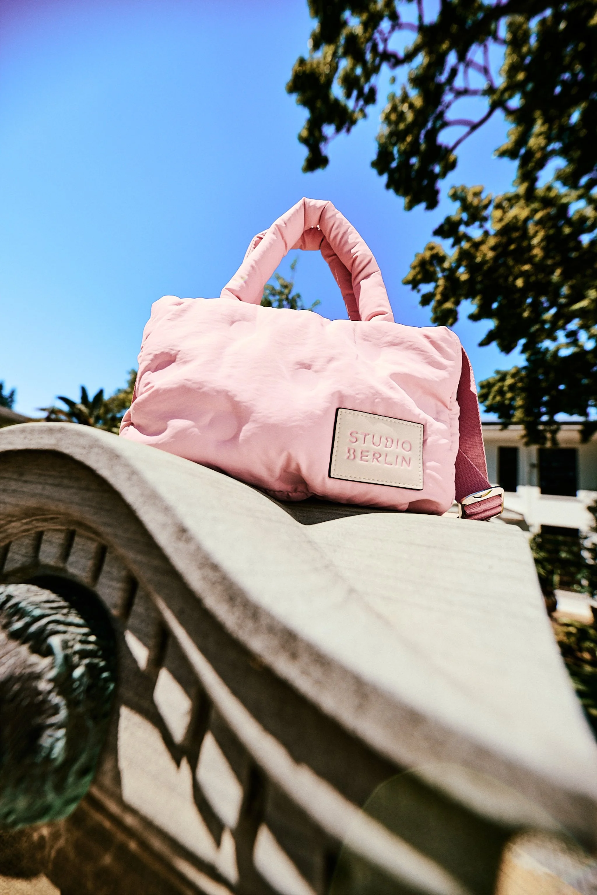 A pink bag labeled "Studio Berlin" resting on a curved wooden surface, with trees and a blue sky in the background.