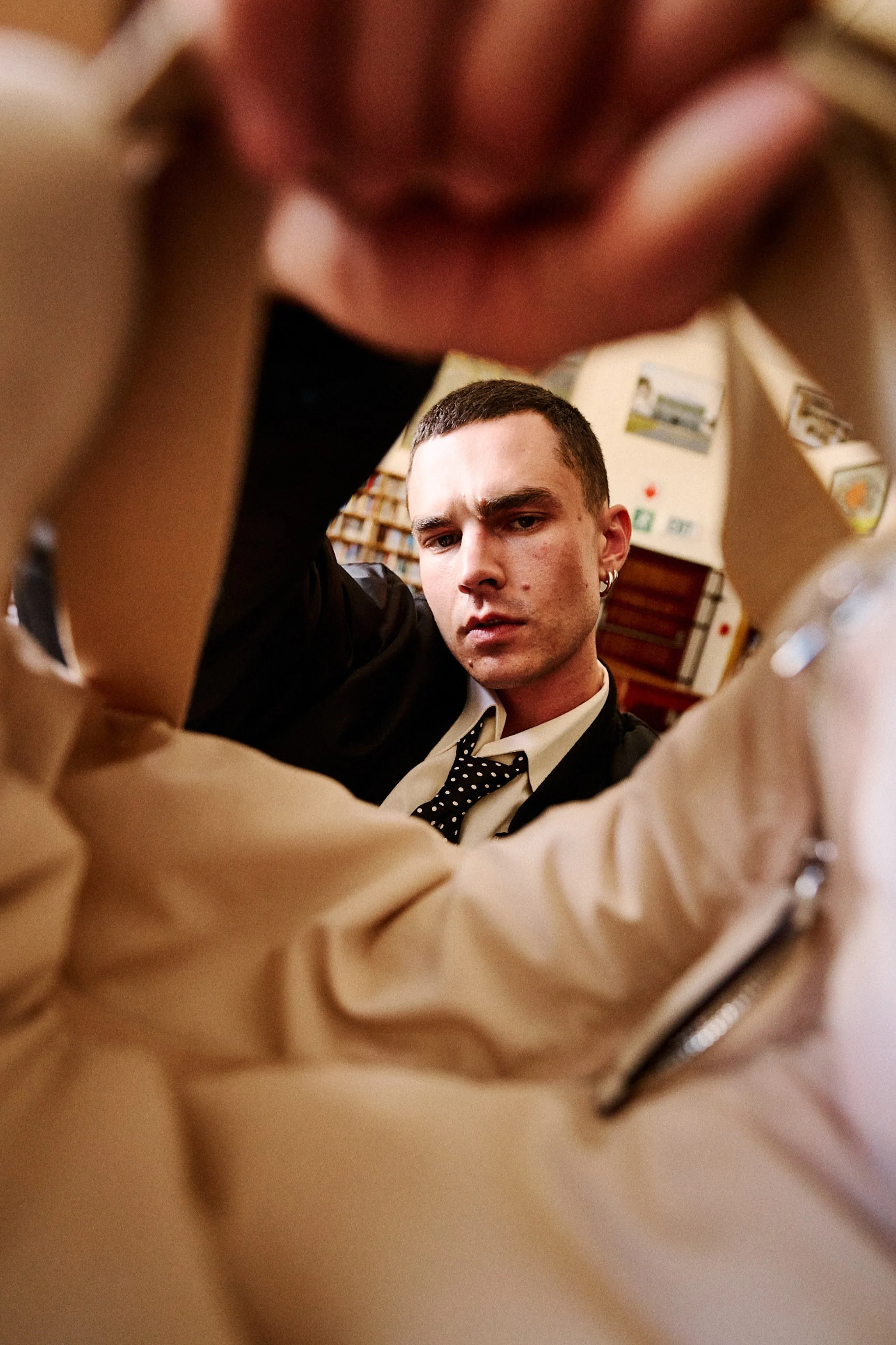 A young man with short dark hair and pierced ears looking directly at the camera, framed by a blurred foreground of a person's knees and thighs, with a background of a bookshelf and posters on the wall.