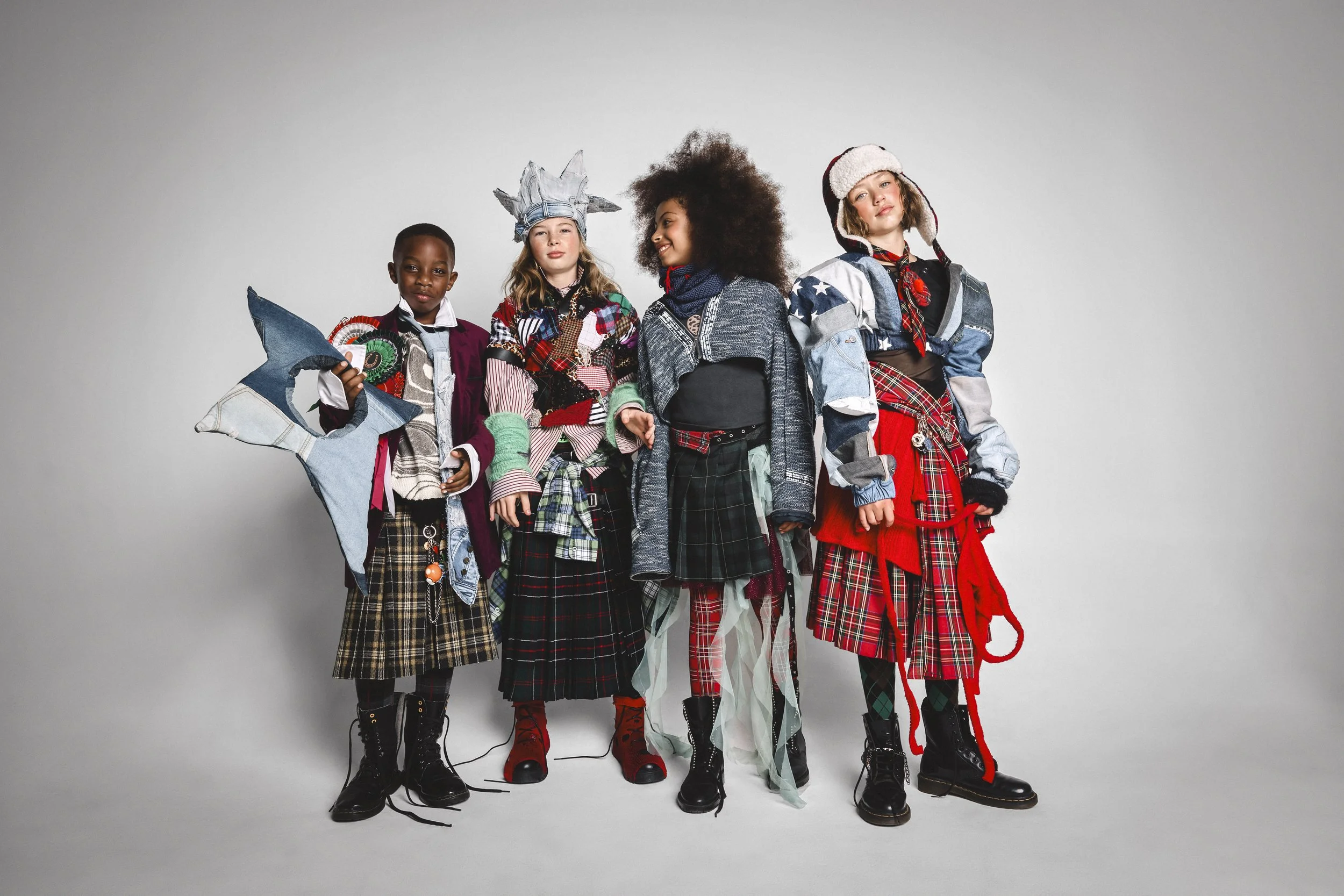 A group of five children standing against a plain gray background, wearing mismatched, layered, and colorful clothing with various accessories, each with a unique expression.