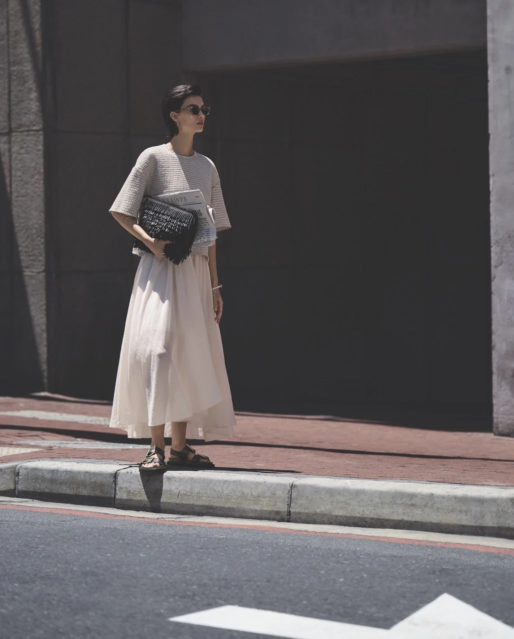 A woman standing on a city sidewalk at a crosswalk. She is wearing sunglasses, a light-colored the, and sandals, holding a black textured clutch and newspapers.