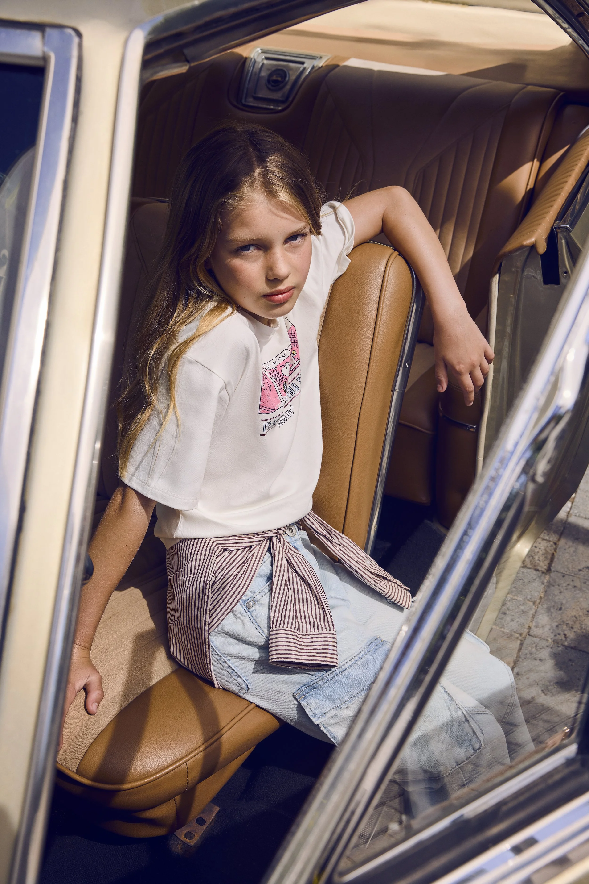 A young girl with long hair looking at the camera while sitting in the back seat of a vintage car with tan leather seats and open door.