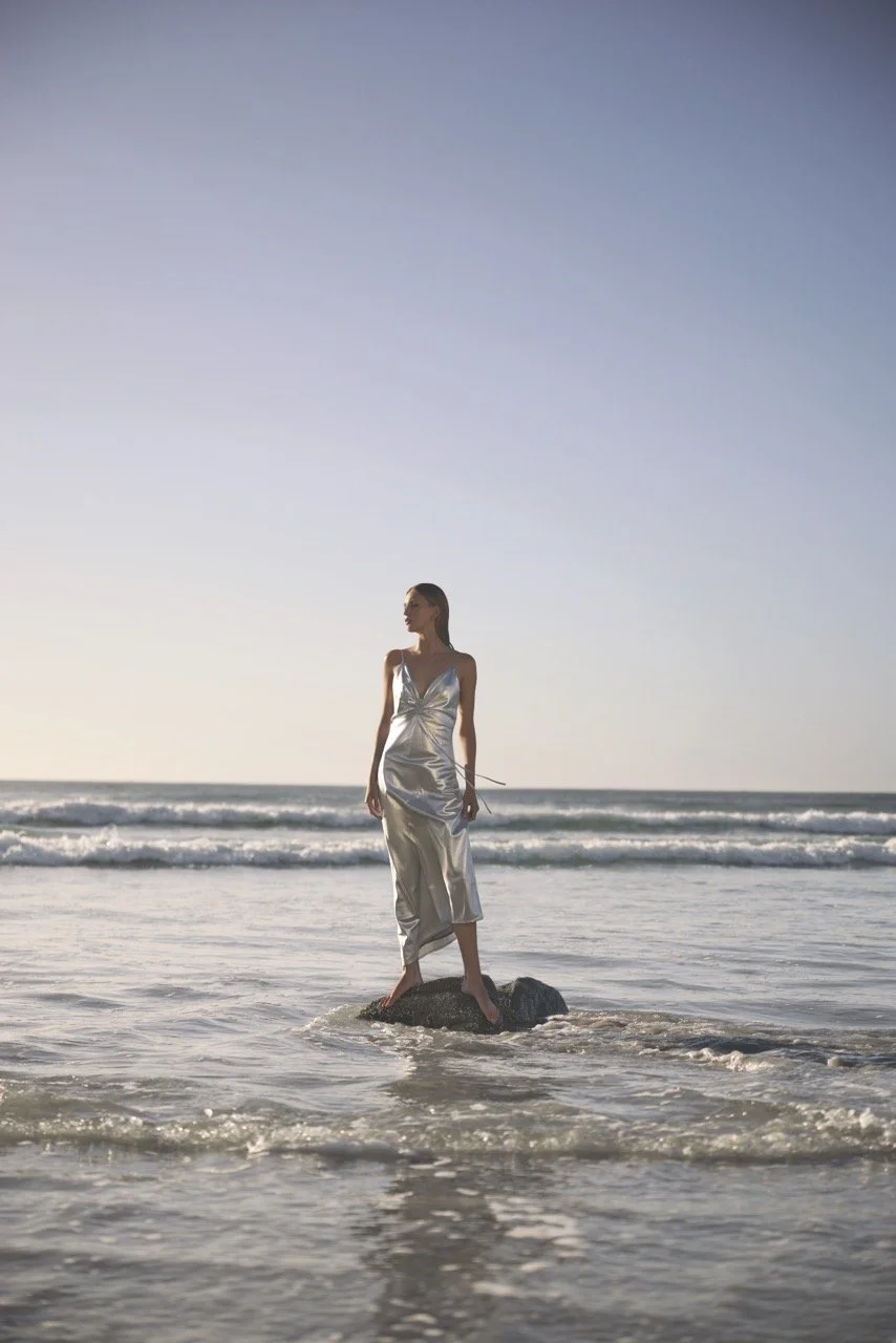 A woman in a silver satin dress standing on a rock in the ocean at sunset.