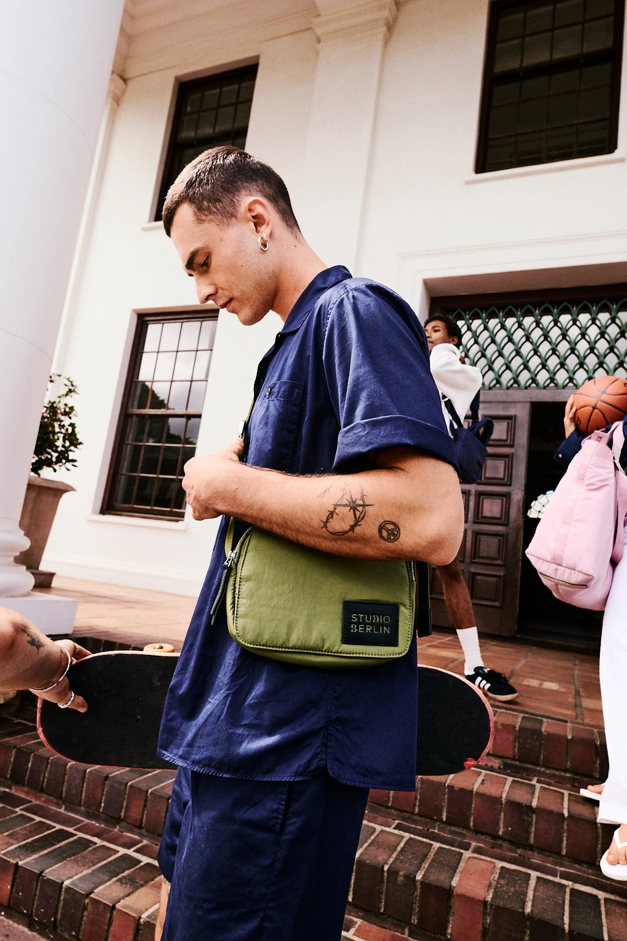 A young man with short dark hair and earrings stands outdoors on brick steps in front of a white building with large windows, wearing a navy blue shirt and carrying a green bag labeled 'Studio Berlin'. Other people are visible in the background, including a person holding a basketball.