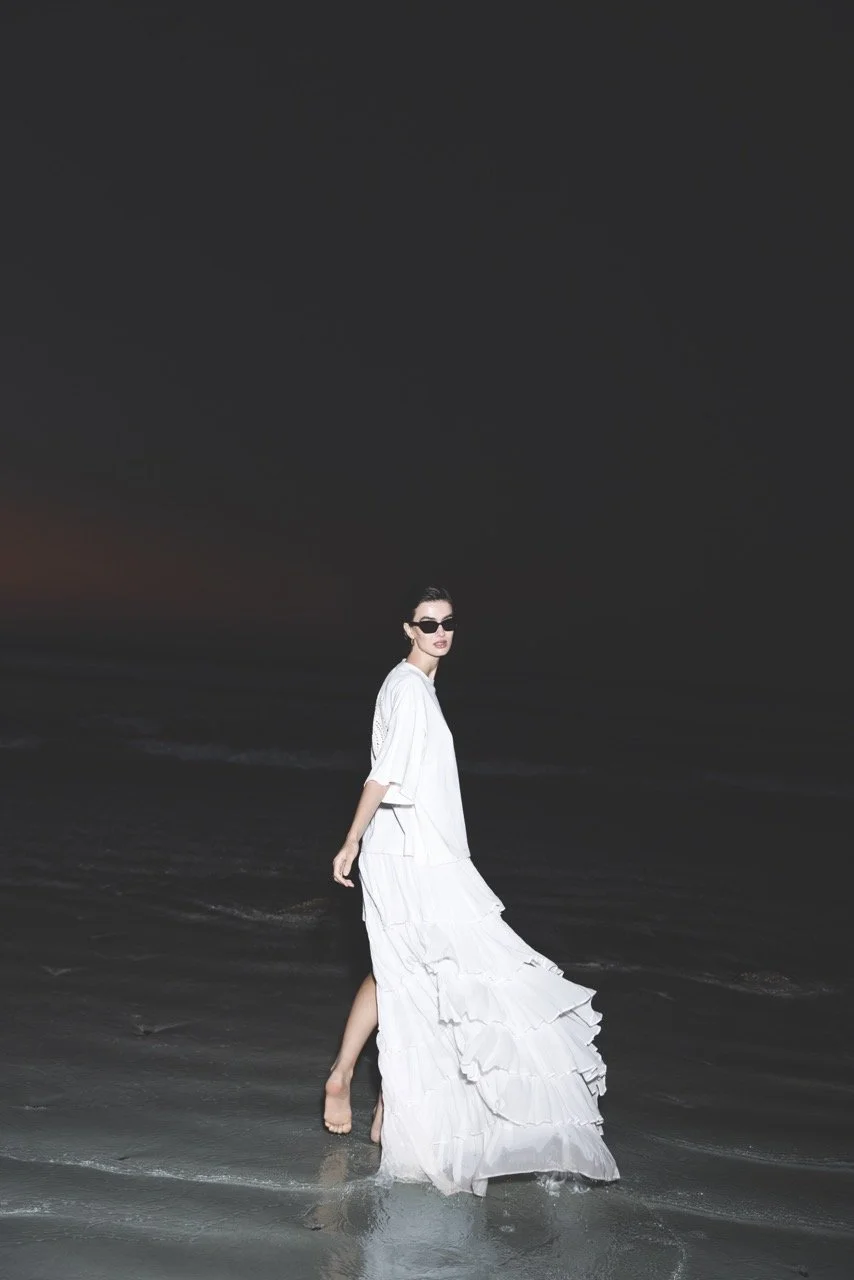 A woman in white dress and sunglasses walking barefoot on the beach at dusk.
