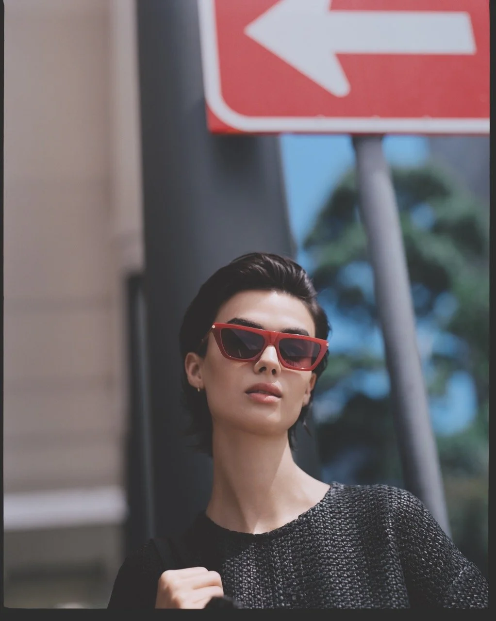 A fashionable woman with short black hair and red sunglasses posing outdoors, with a street sign and trees in the background.