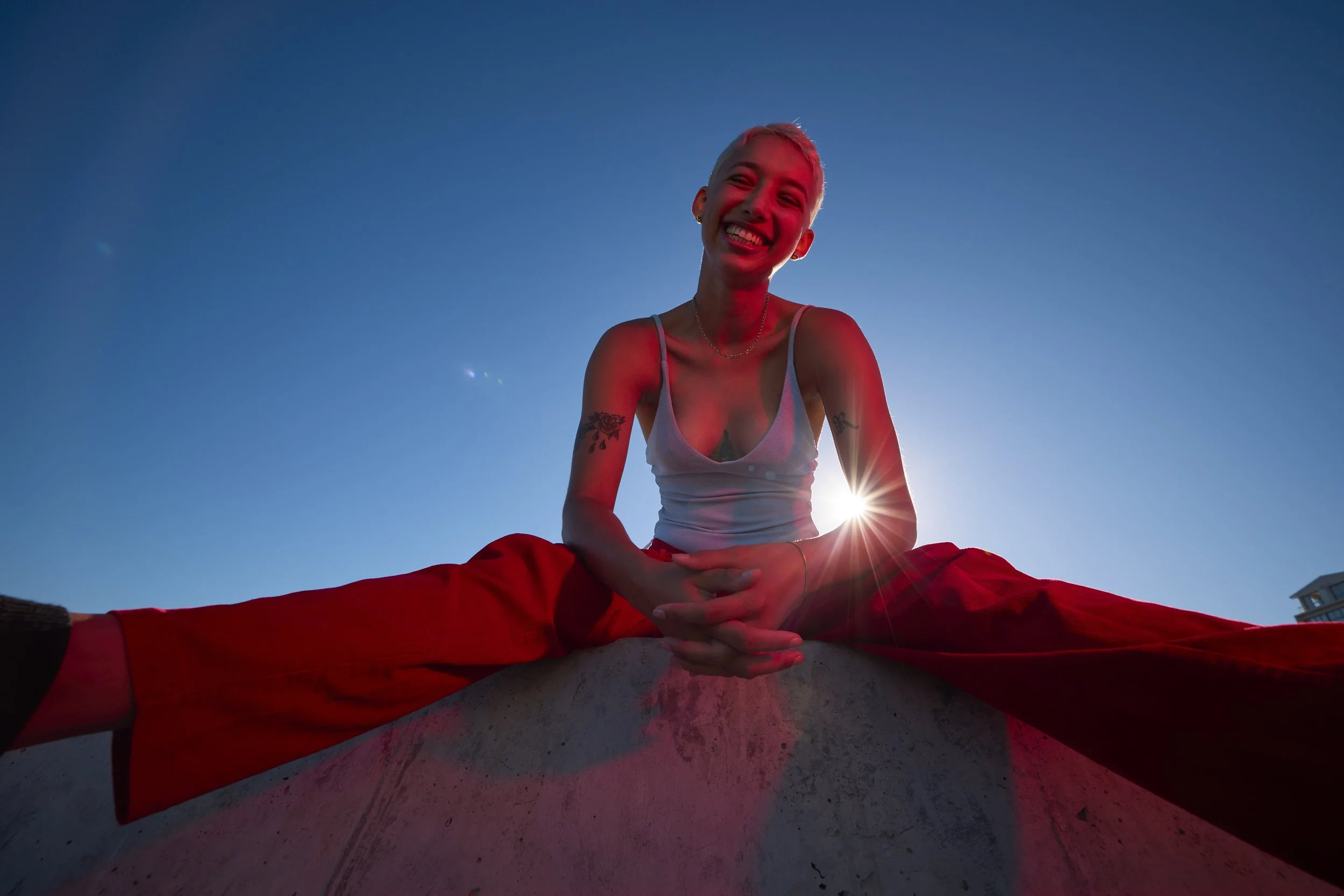 A woman with short hair and tattoos smiling and sitting cross-legged on a yoga mat outdoors during sunset, with the sun behind her. She is wearing a white tank top and red pants.