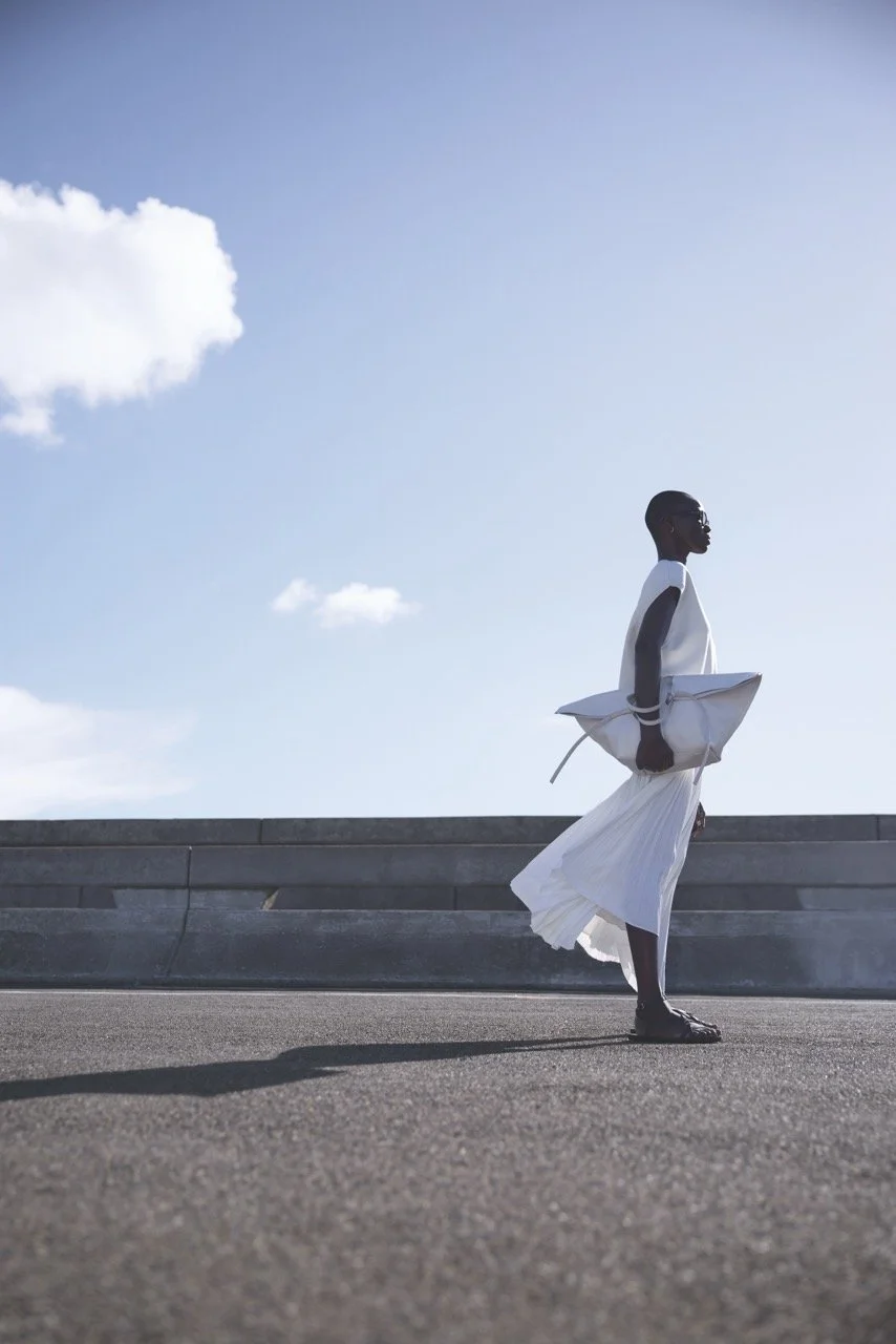 A person walking outdoor under a blue sky with some clouds, carrying a large white bag and dressed in white clothing.