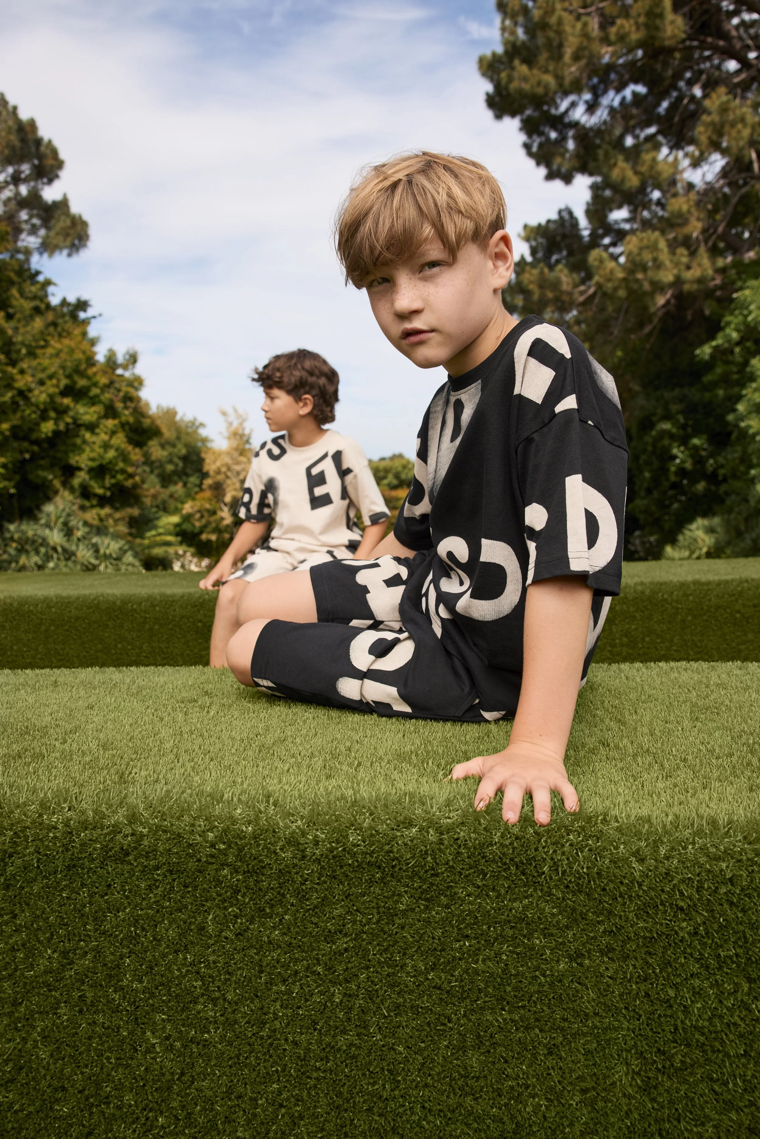 Two boys sitting on the grass outdoors, one in the foreground with a serious expression, the other in the background looking away. Trees and a partly cloudy sky are in the background.