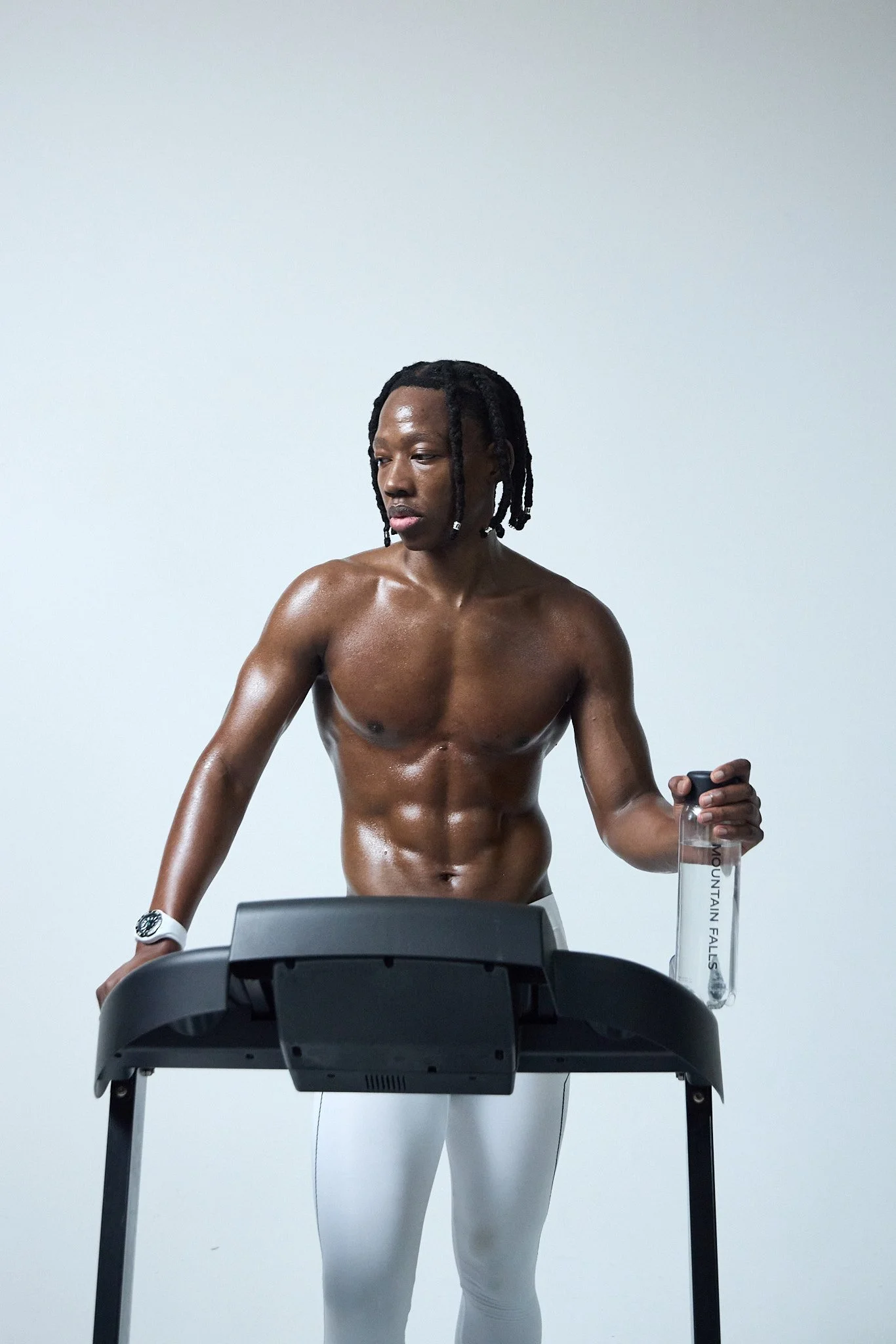 A shirtless man with dreadlocks is walking on a treadmill, holding a water bottle in his left hand, with a neutral facial expression, in front of a plain light-colored wall.