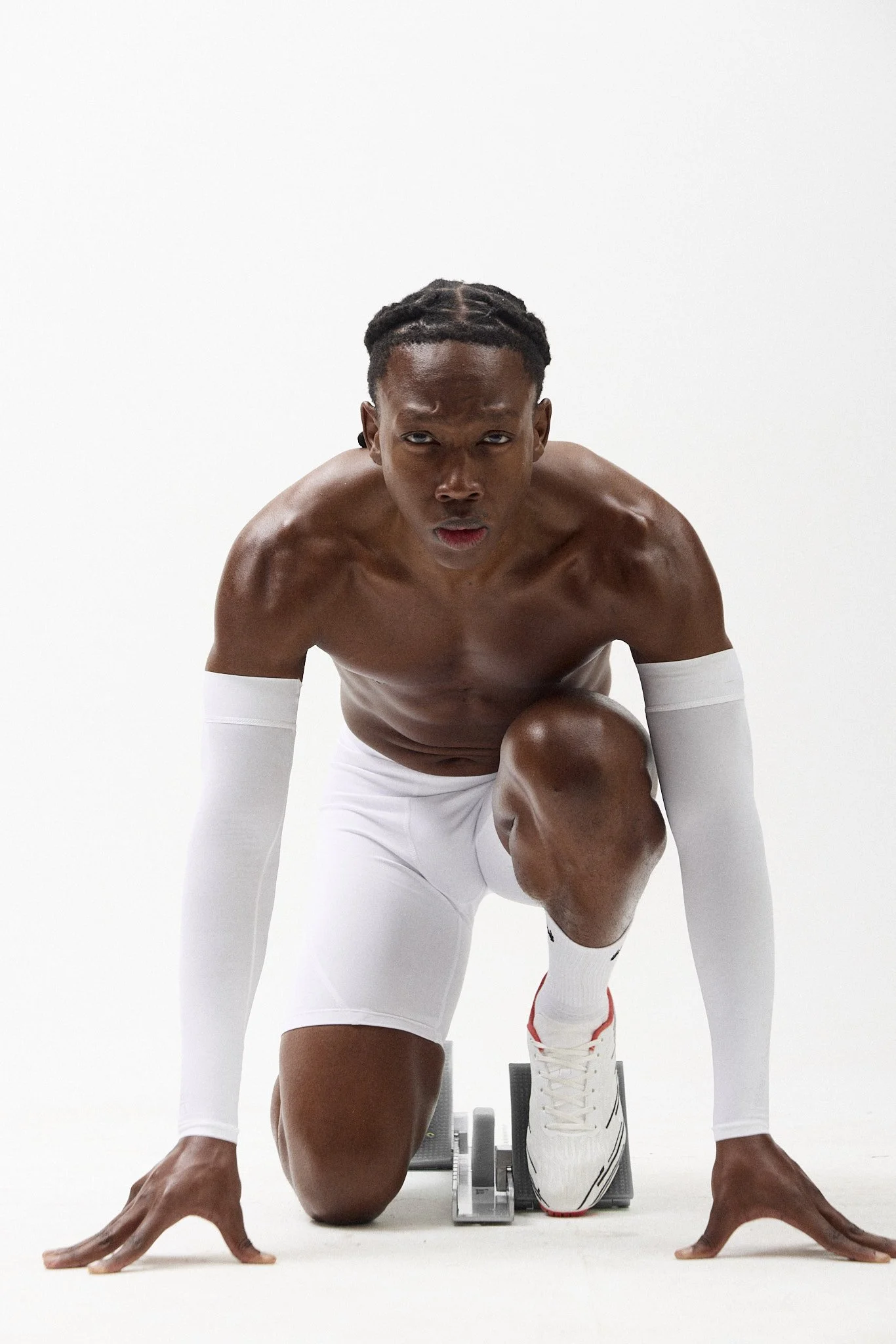 A shirtless man in white athletic gear, including long sleeves, shorts, and sneakers, poses on a treadmill against a plain white background, with a serious expression and hand on the floor.