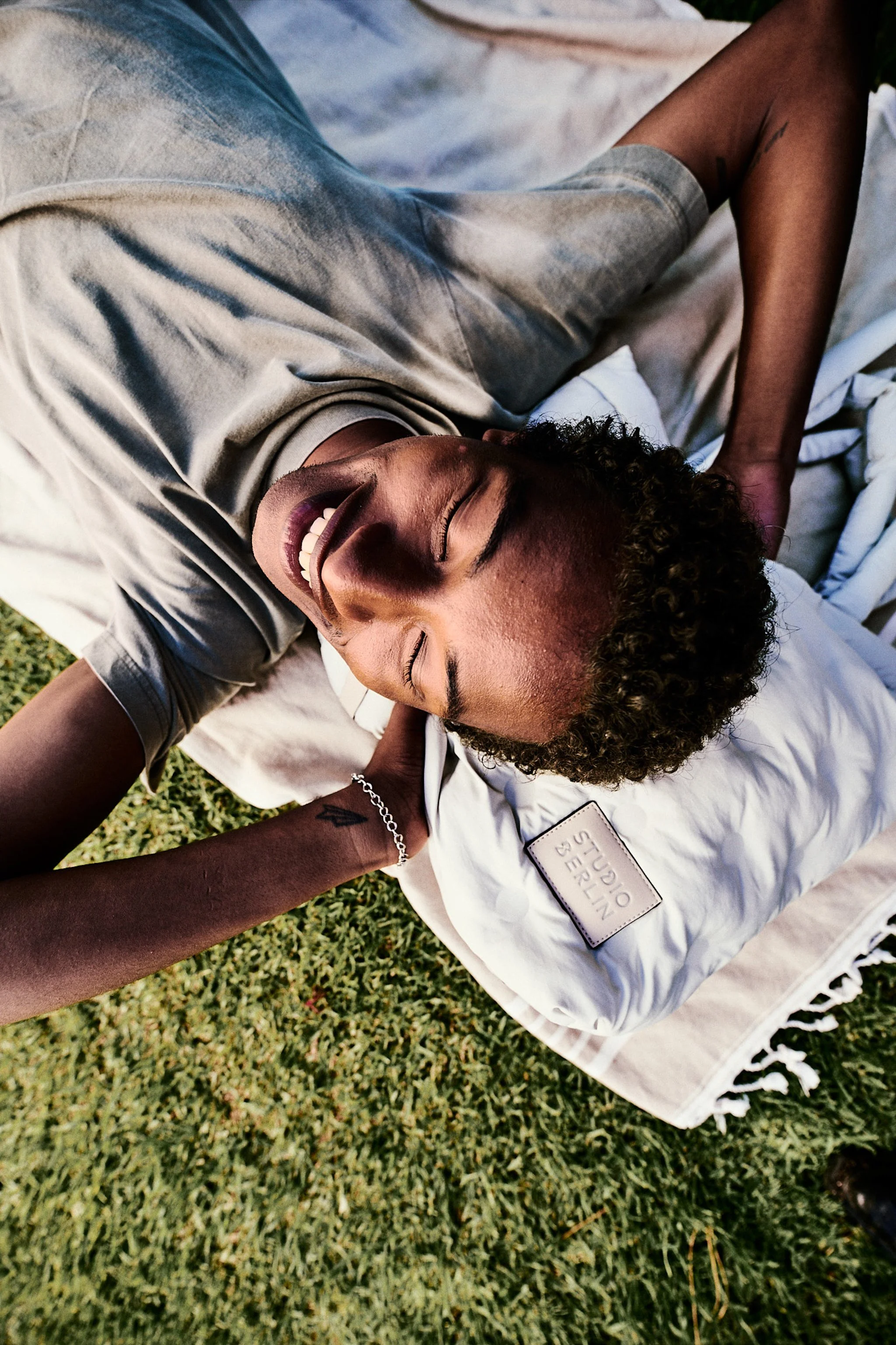 A person with curly hair lying on a white blanket on the grass, smiling with eyes closed. They are wearing a grey t-shirt and beige shorts. One arm is bent behind their head and the other is extended across the blanket. The person has a silver bracelet and a tattoo on their wrist.