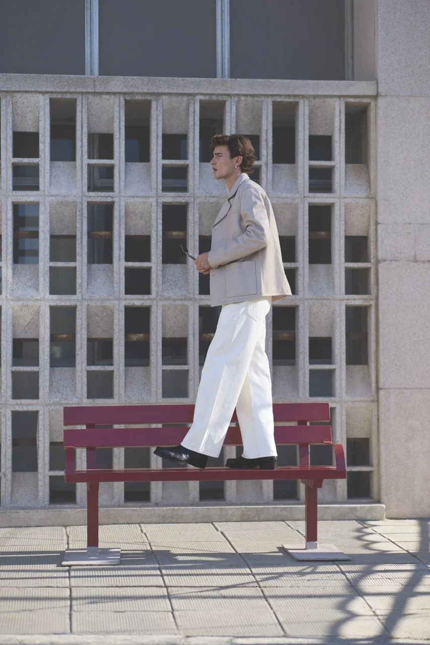 Person walking on a red park bench, holding sunglasses, in front of a concrete building with patterned windows.