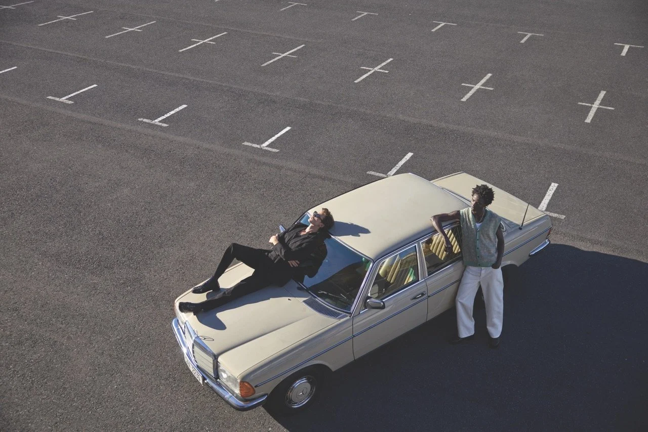 A woman is lying on the hood of a beige vintage car, with her head resting back and eyes closed. A man stands beside the car, leaning against the door with arms crossed, looking to the side. The scene is in an empty parking lot with marked parking spaces.
