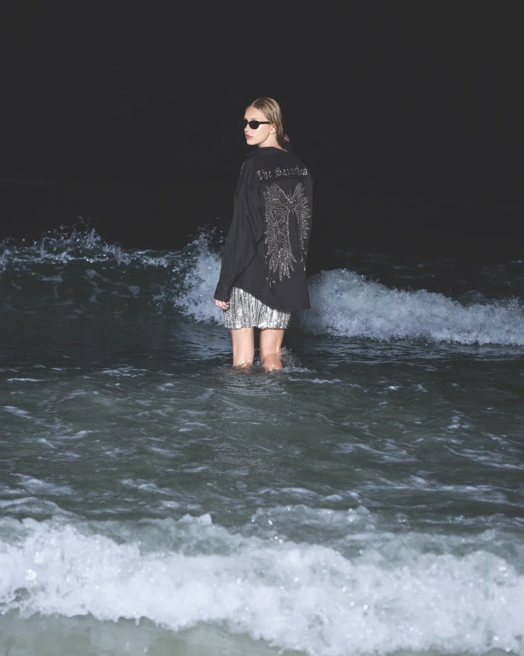 A woman standing in shallow ocean water wearing sunglasses, a black jacket with a large angel wings design on the back, and a sparkly skirt during nighttime.