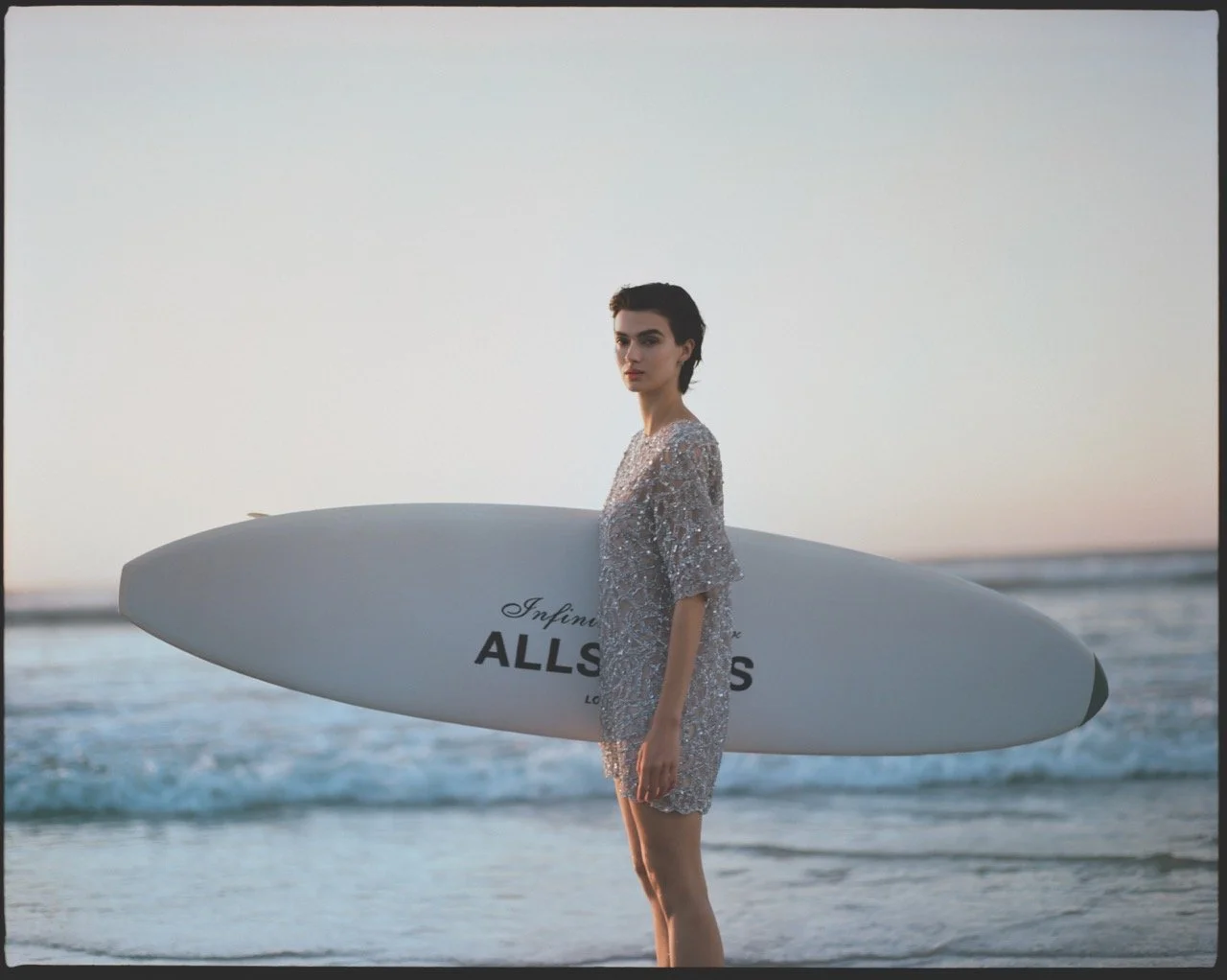 A woman in a shiny, sequined dress holding a surfboard on the beach at sunset.