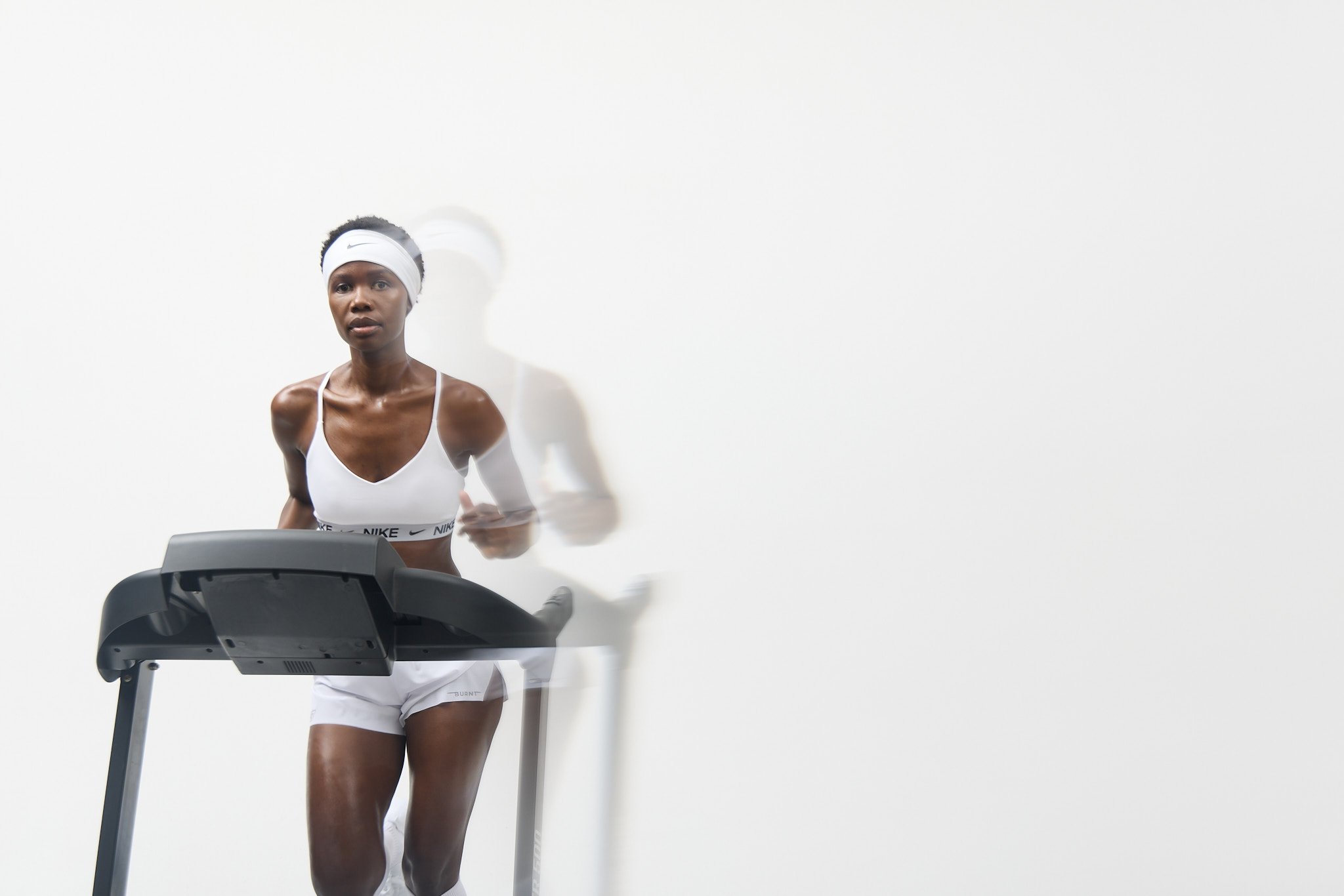 A woman running on a treadmill, dressed in white athletic wear and a headband, in a minimal white room.