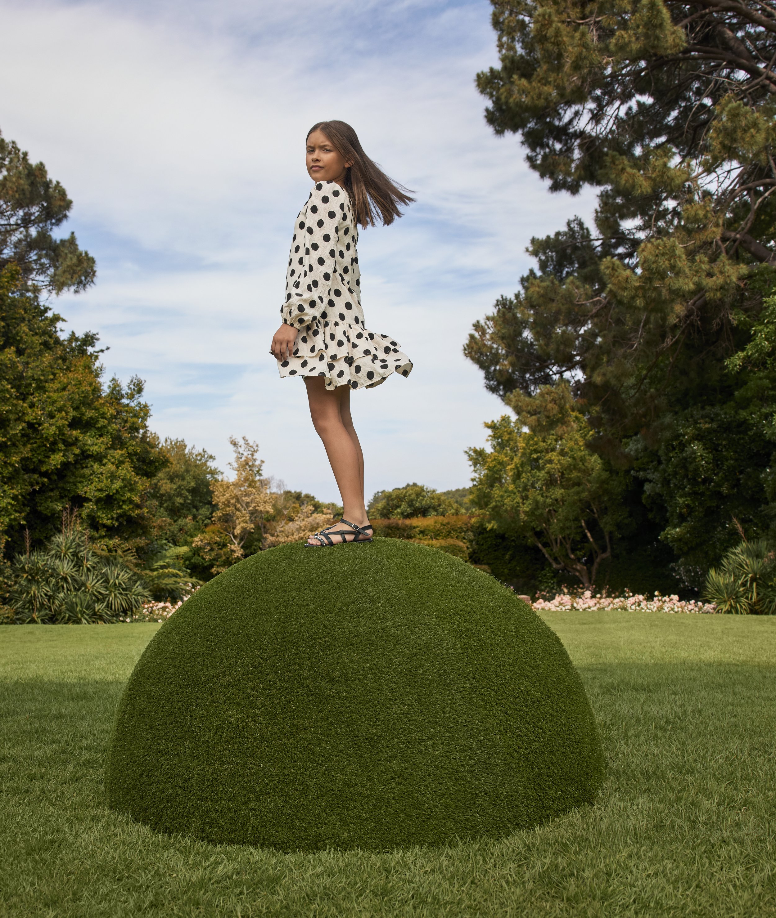 A young girl in a polka dot dress standing on a round, black, turf-covered mound in a lush green park with trees and flowers in the background, under a partly cloudy sky.