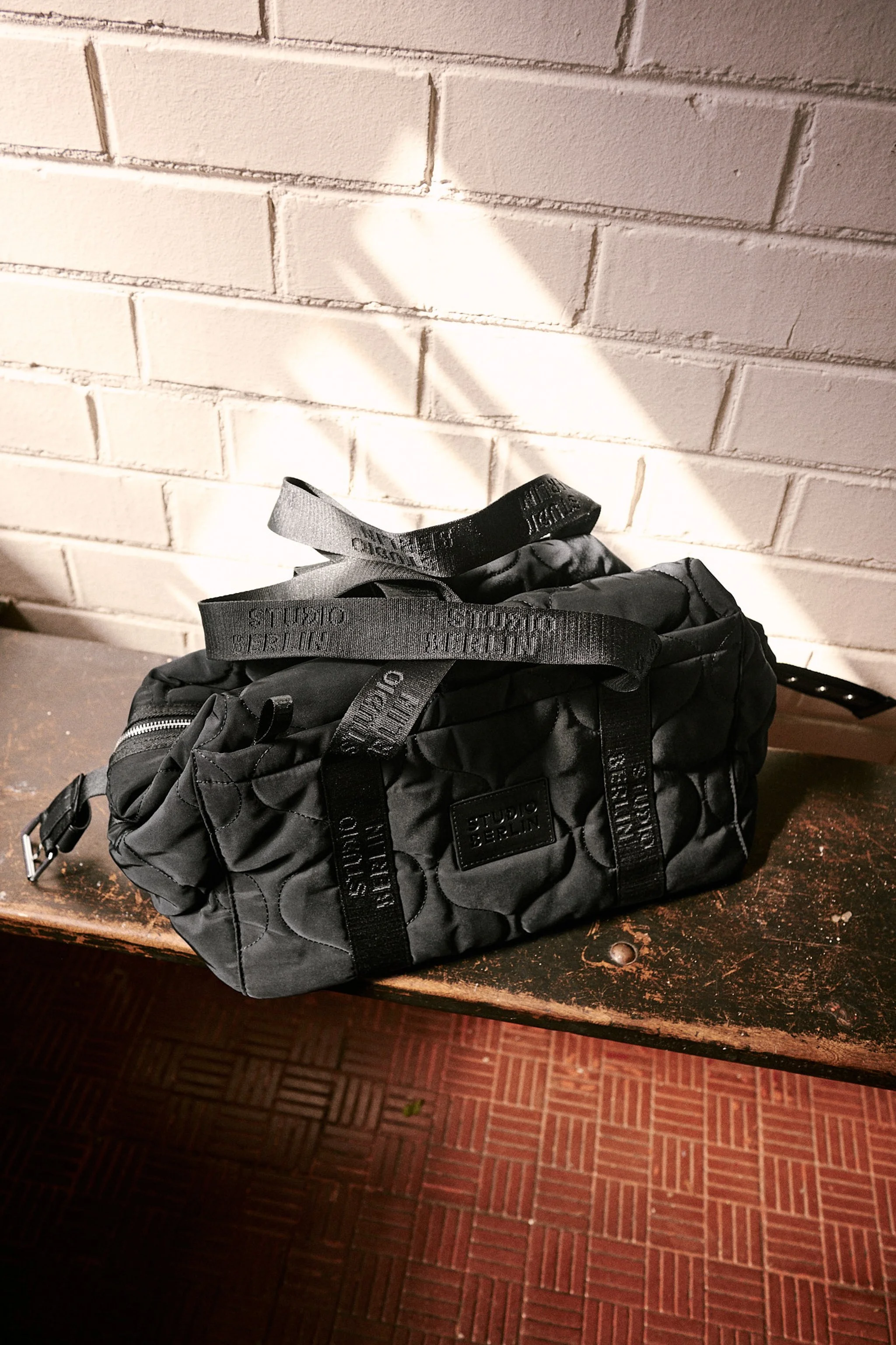 A black quilted backpack with straps that have text on them, placed on a wooden table against a white brick wall. Light from a window cast diagonal shadows on the wall.