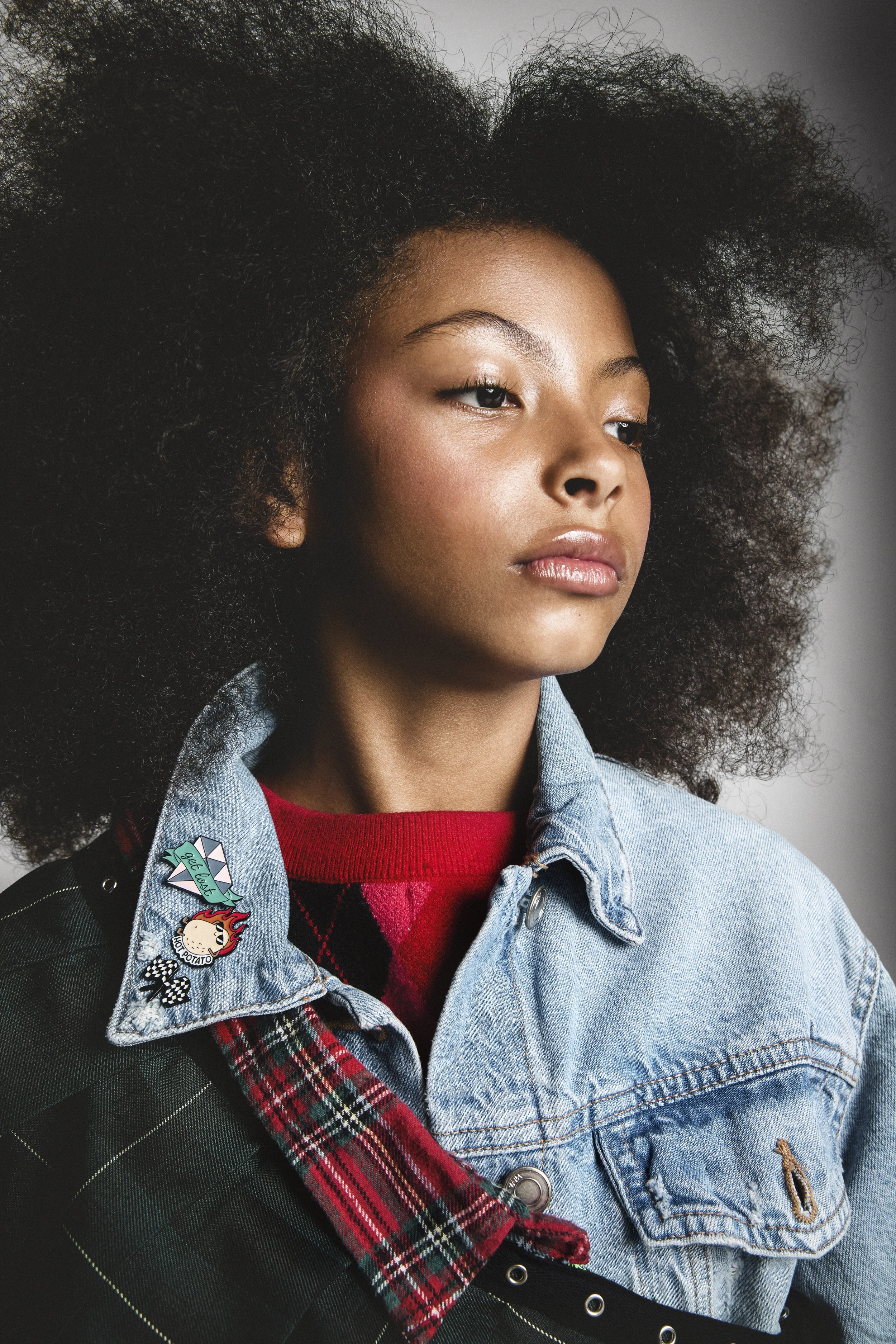 A young woman with voluminous natural curly hair, wearing a denim jacket with various patches, a red sweater underneath, and a plaid accessory draped over her shoulder.