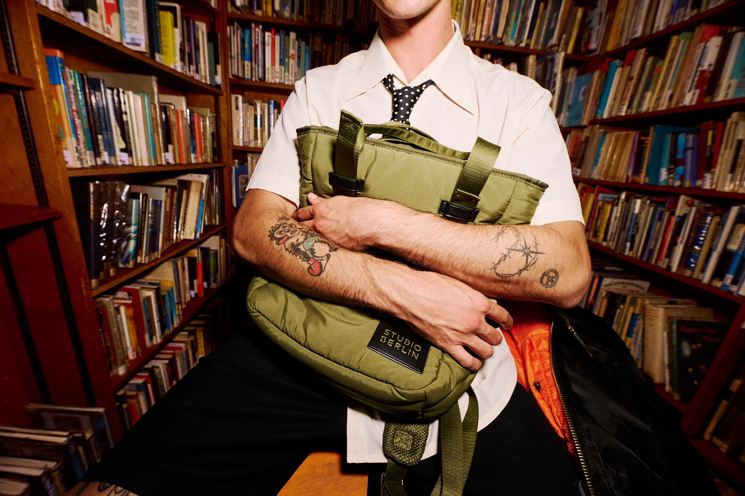 Person in a library holding a green padded bag, wearing a white shirt and tie, with tattoos on their arms, surrounded by shelves of books.