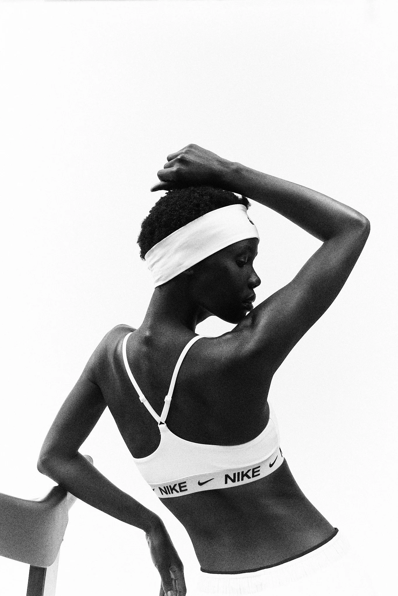 Black and white image of a woman wearing a white Nike sports bra, white shorts, and a headband, with her left arm raised and her right arm resting on a mannequin torso.