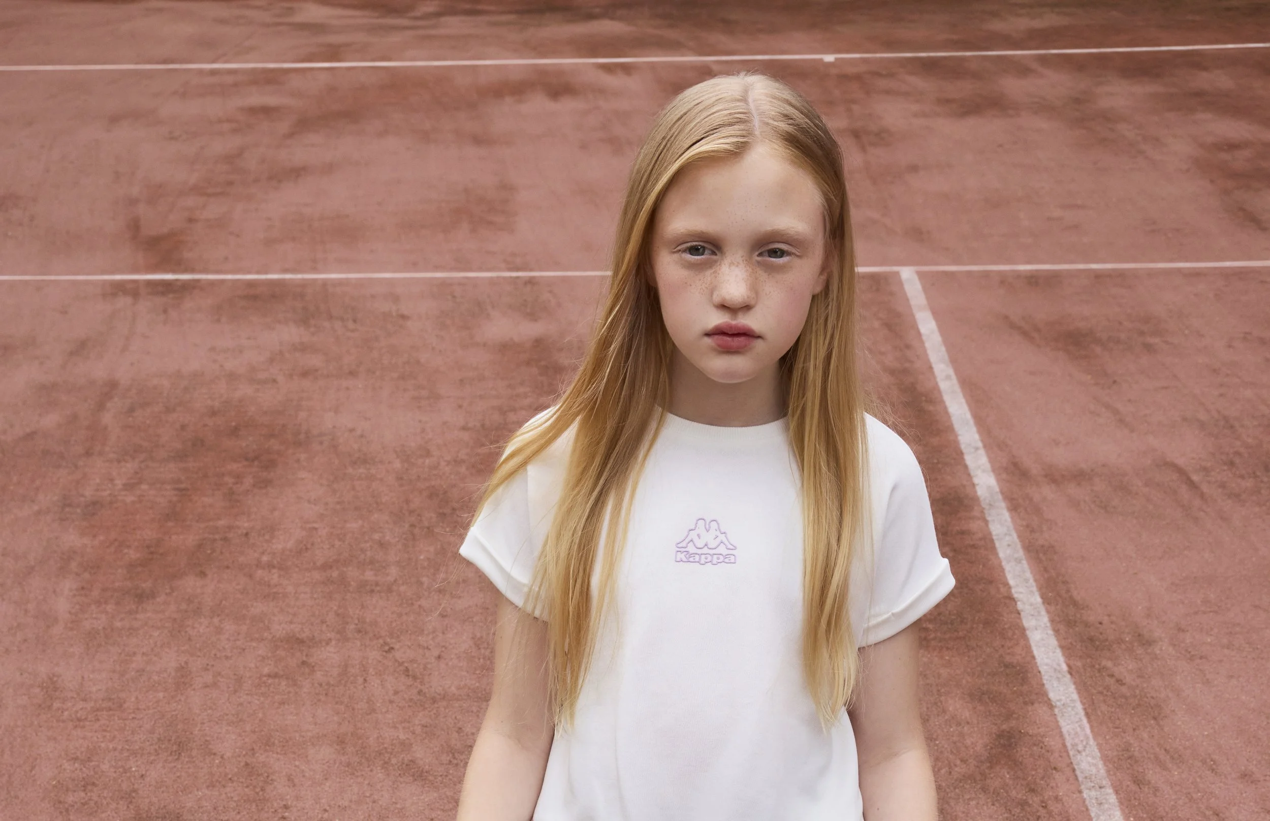 A young girl with long blonde hair and freckles standing on a reddish outdoor clay tennis court.