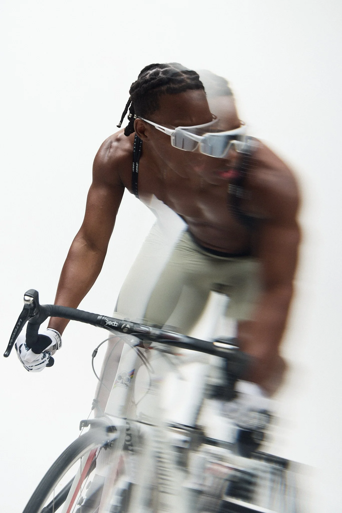 A male cyclist wearing sunglasses and gloves riding a bicycle indoors with a minimalistic white background.