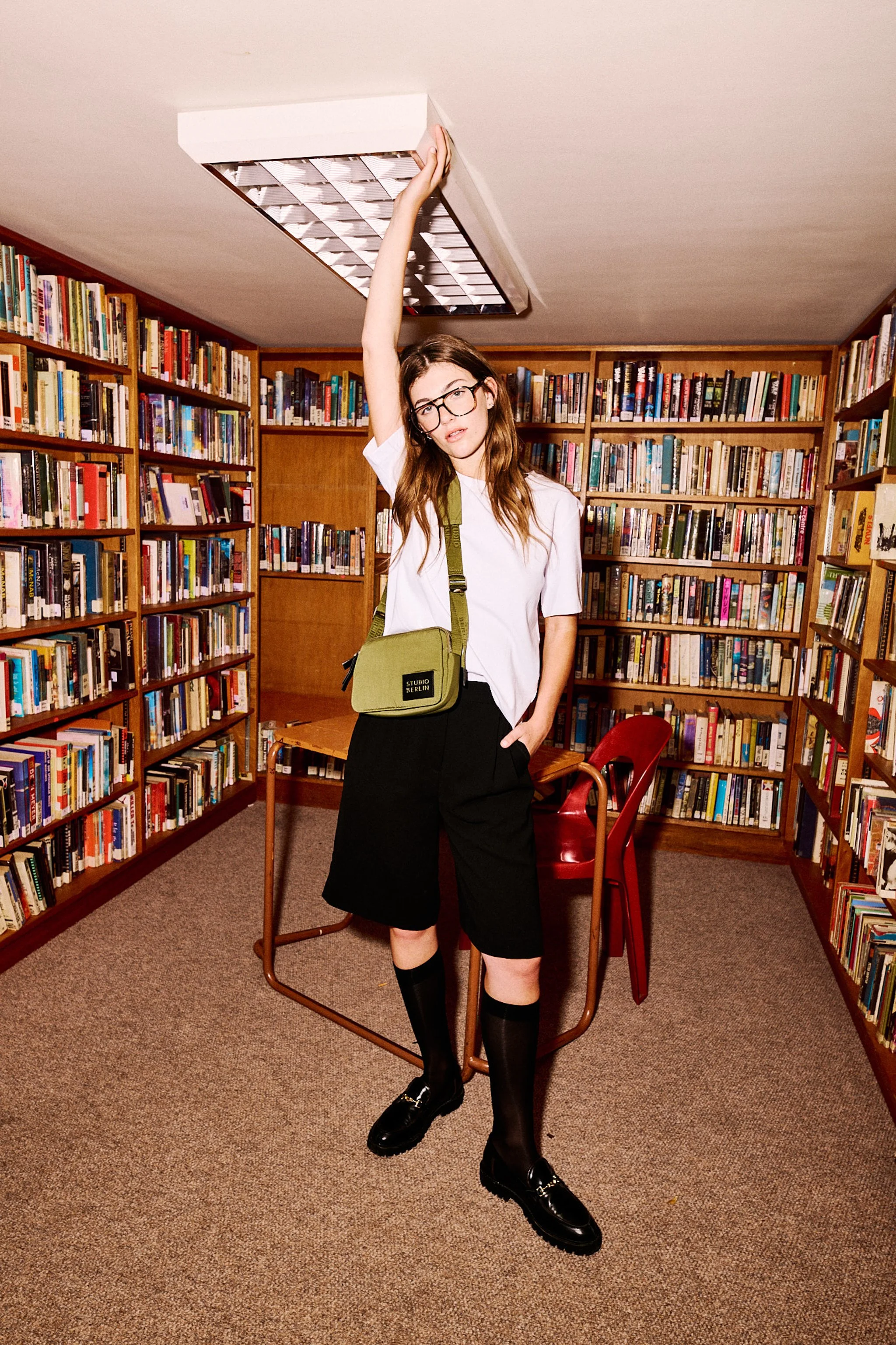 A young woman with long brown hair, glasses, and light skin stands in a library. She is wearing a white t-shirt, black knee-high socks, black shoes, and black shorts. She has a green shoulder bag and is holding onto a ceiling light fixture with her right hand, while her left hand is in her pocket. The background shows wooden bookshelves filled with books.