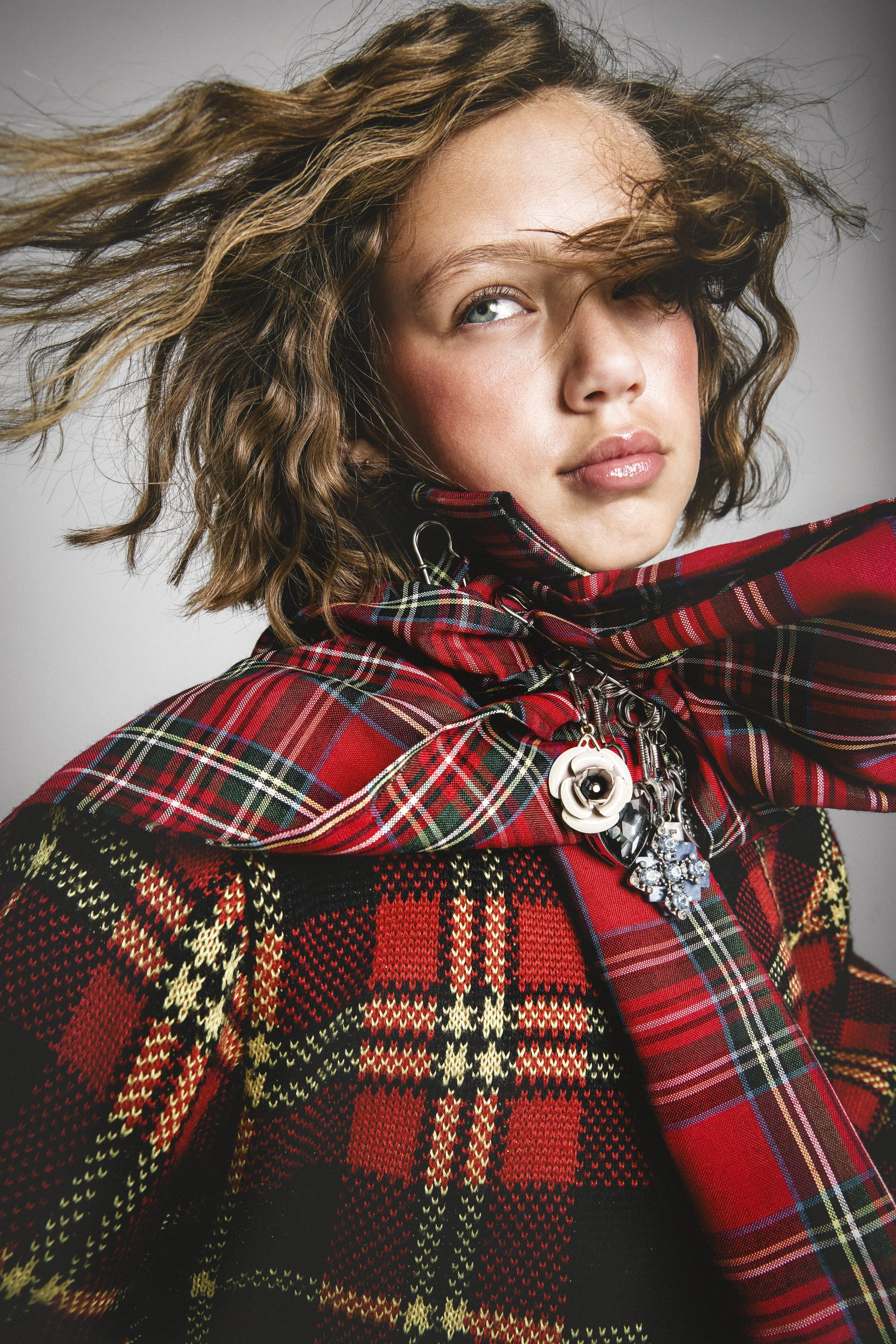 A woman with wavy hair wearing a red tartan scarf and a holiday sweater with a Christmas plaid pattern, complemented by a necklace with a flower-shaped pendant and large jewelry charms.