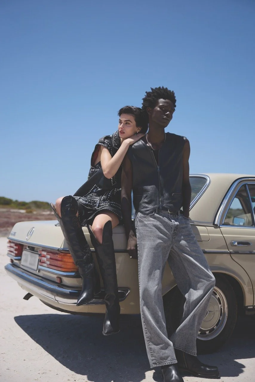Two fashionably dressed people with dark and light skin tones leaning against a beige vintage Mercedes-Benz car in a barren landscape under a clear blue sky.
