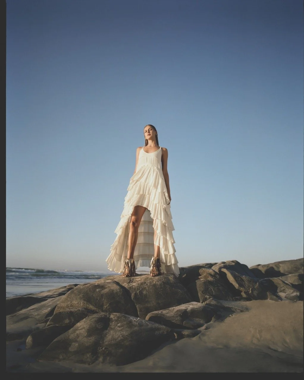 A woman wearing a long, flowy white dress standing on rocks on the beach with the ocean and a clear blue sky in the background.